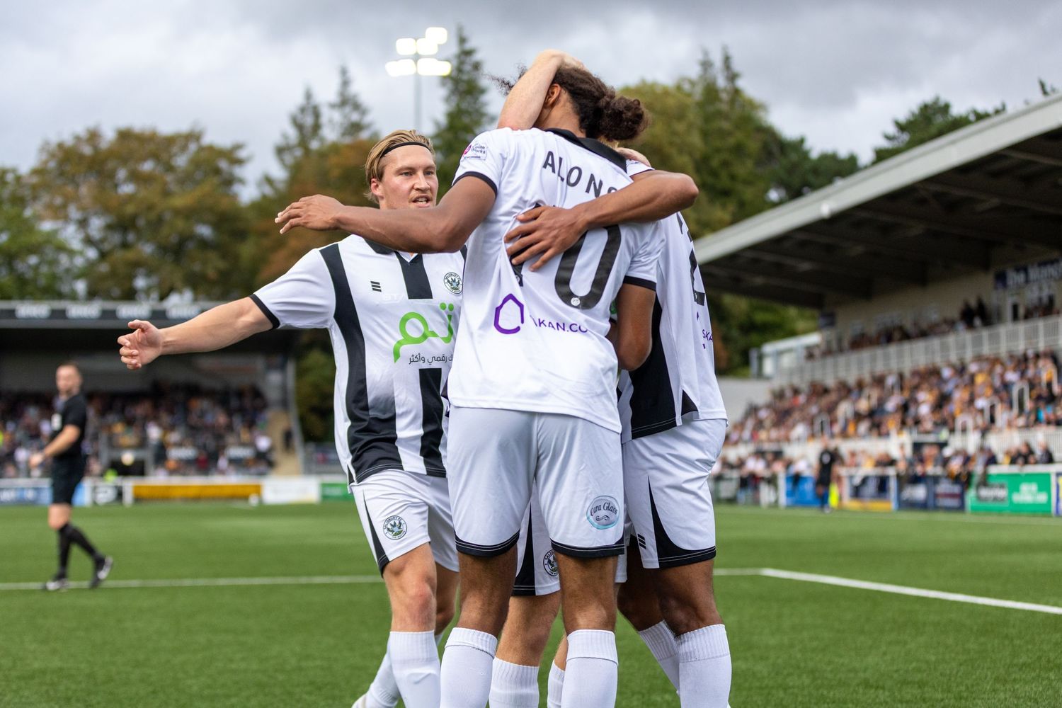 Football players in white and black kits celebrating a goal together on a sunny match day at a packed stadium.