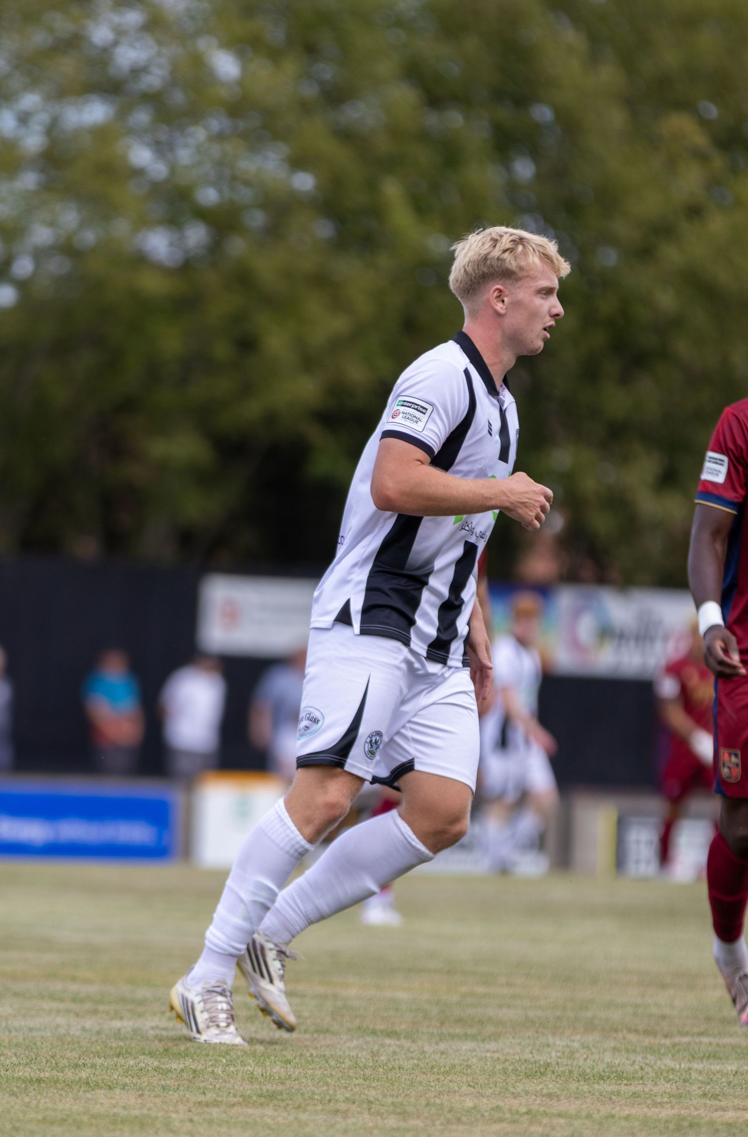 A blonde footballer in a black and white striped kit runs across a grass pitch during a competitive match.