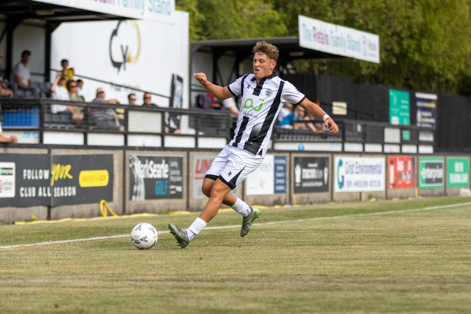 Young footballer in black and white striped kit controls the ball during a sunny outdoor match with spectators watching.