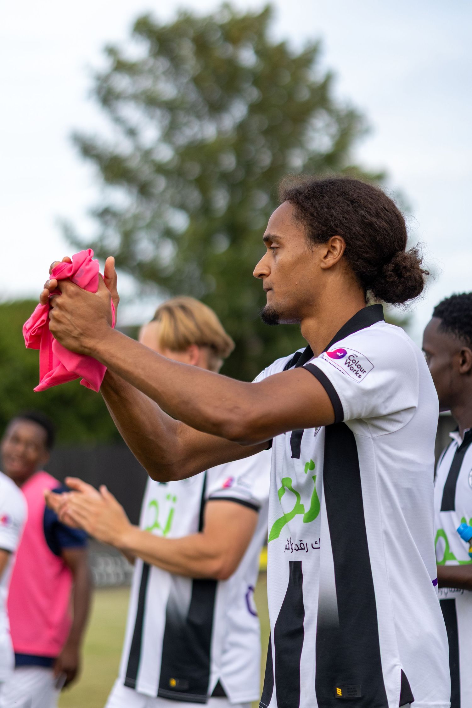 Football player in black and white strip holds pink cloth while teammates celebrate nearby.