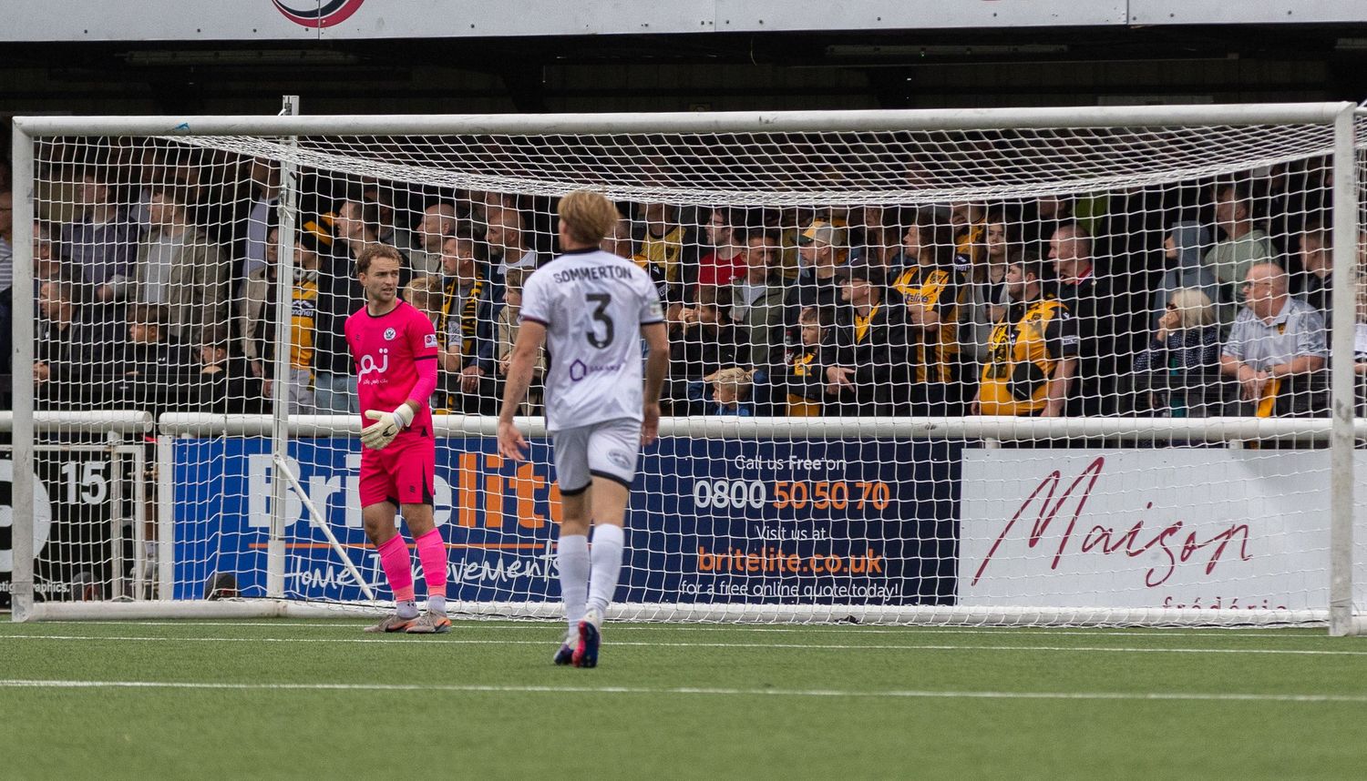 Football players stand near the goal net during a match, with fans watching from the stands in the background.