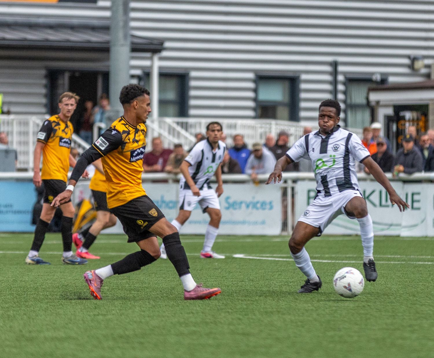 Two soccer players compete for the ball during a match on a green pitch, with teammates and spectators visible in the background.