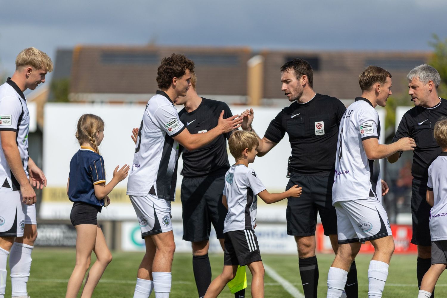 Soccer players in white uniforms shake hands with a referee and young mascots before a match on a sunny day.