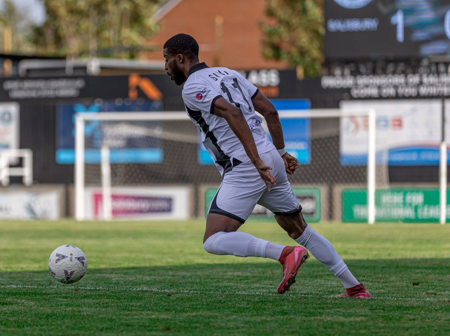Soccer player in white kit dribbling a ball on a green field during a sunny outdoor match.