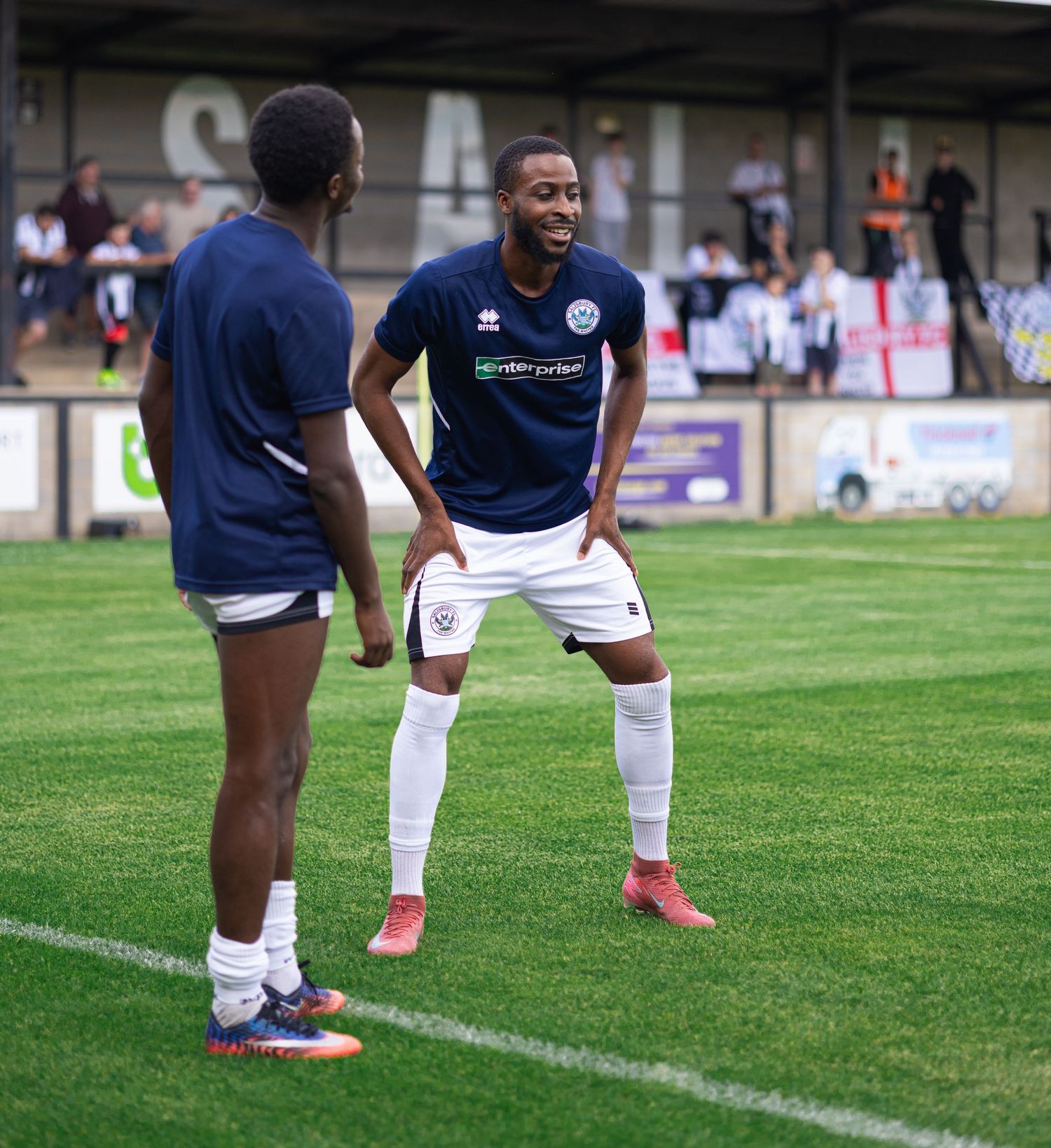 Two soccer players in navy blue uniforms chat on a green pitch during a pre-match warmup at an outdoor stadium.
