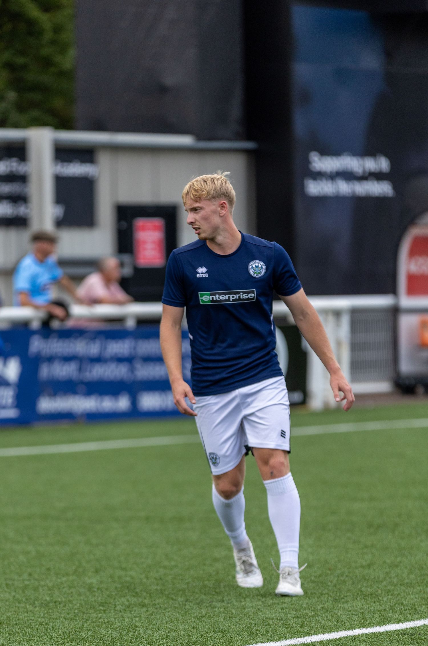 Young footballer in navy Enterprise-sponsored kit walks on green pitch during a sunny outdoor match.