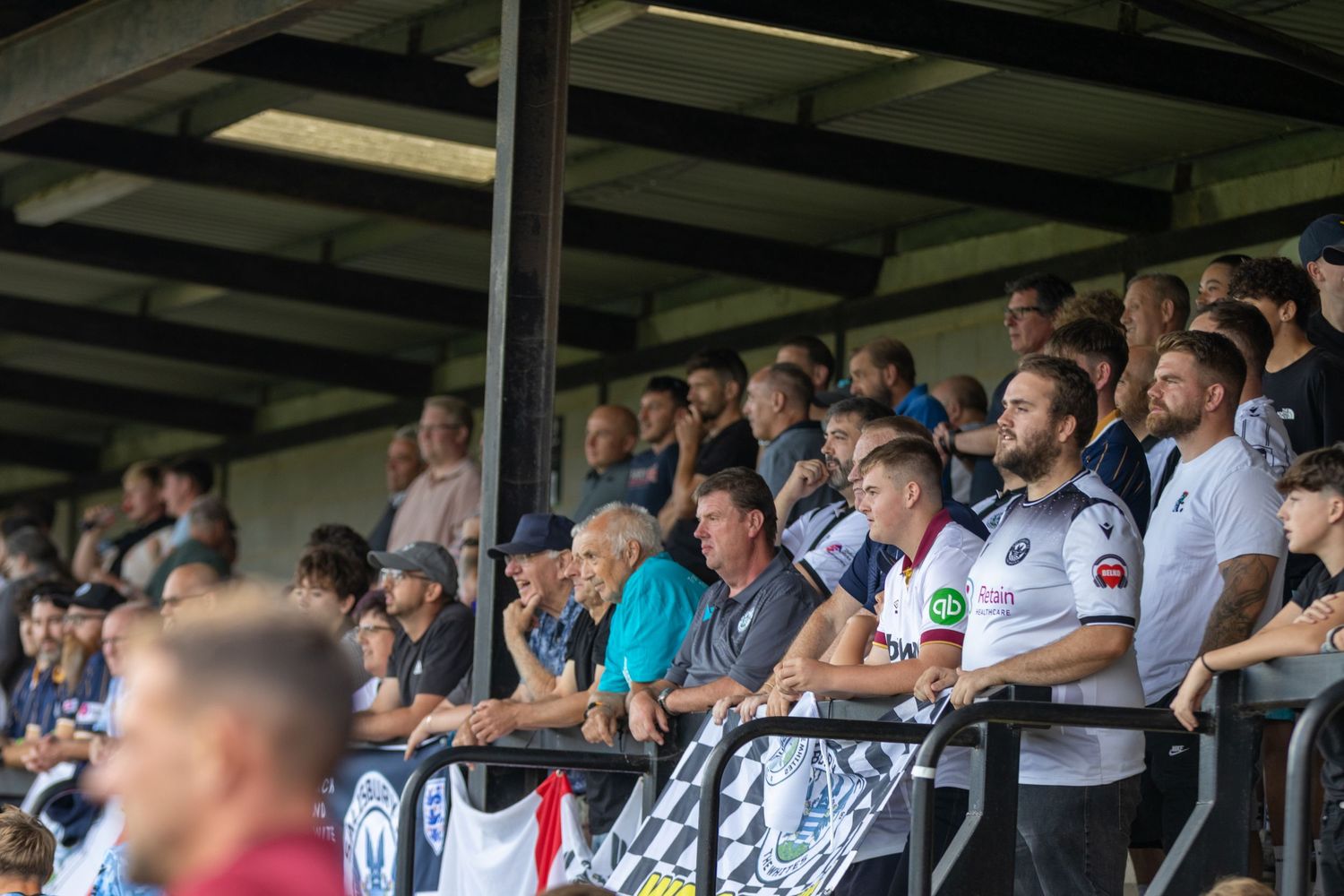 Fans watching a football match from a covered stand at a non-league soccer ground on a sunny day.