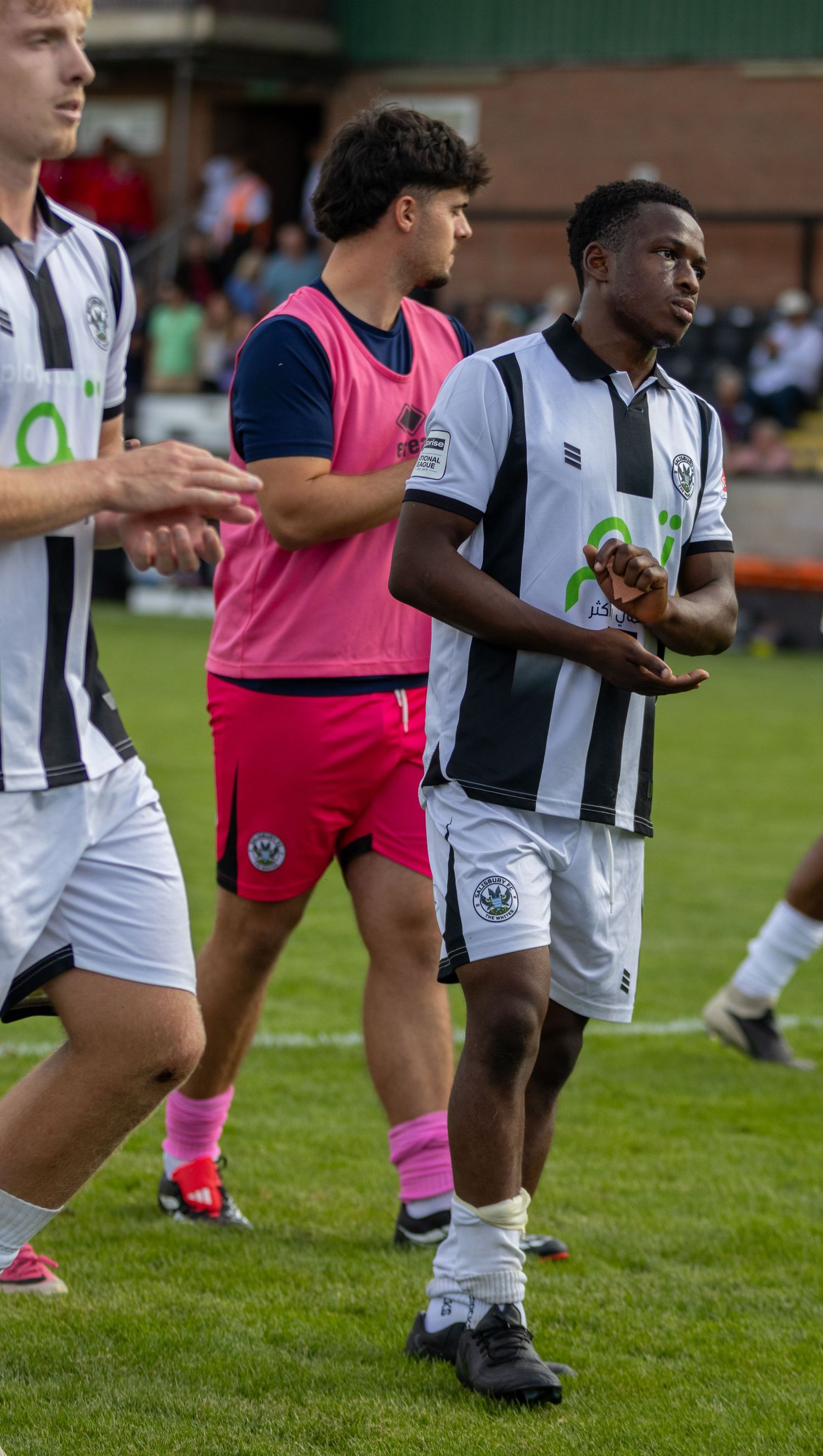 football players in black and white striped uniforms walk on a green field, with one player wearing a pink training bib.