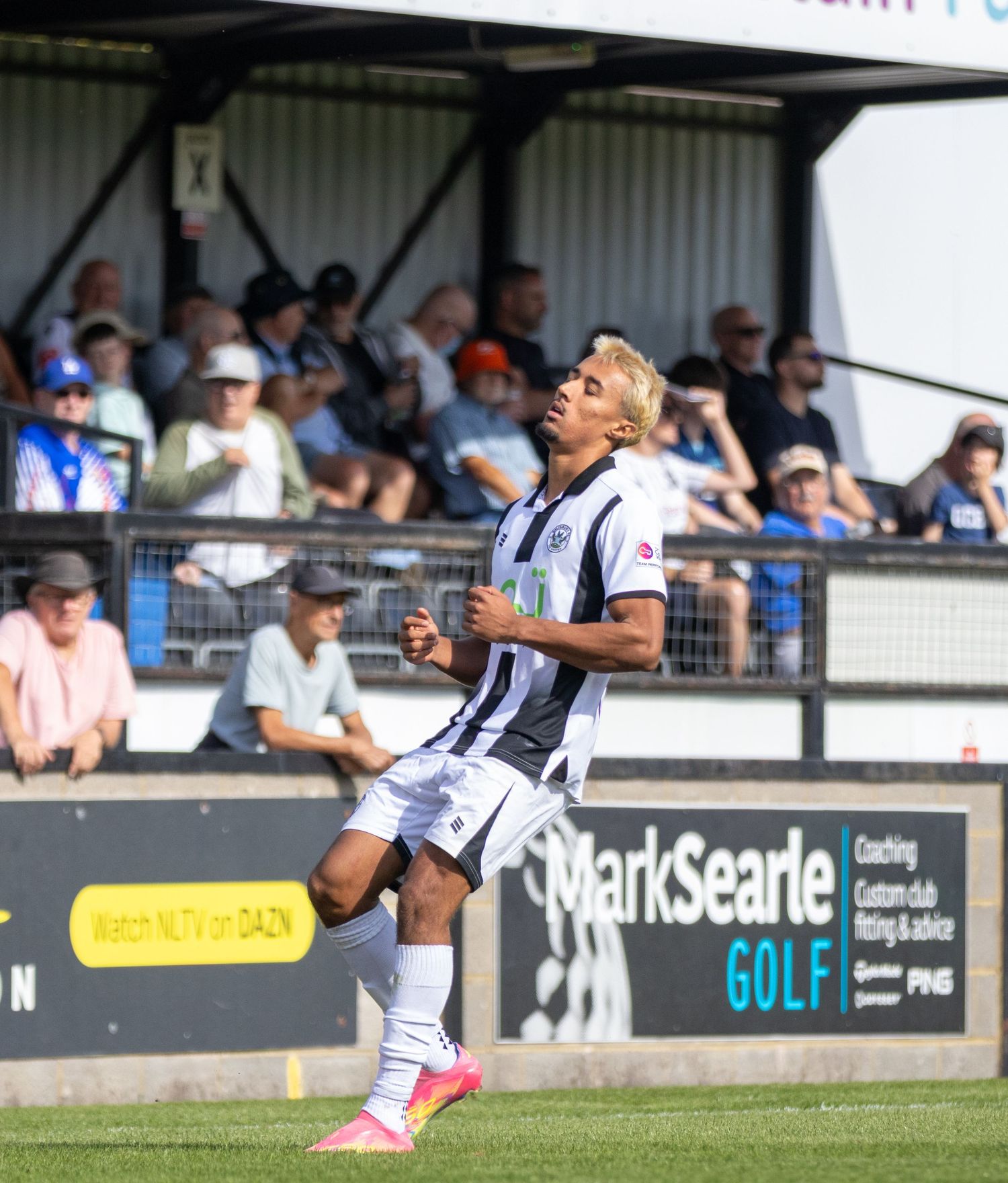 Footballer in black and white striped kit celebrates near Mark Searle Golf advertising board.