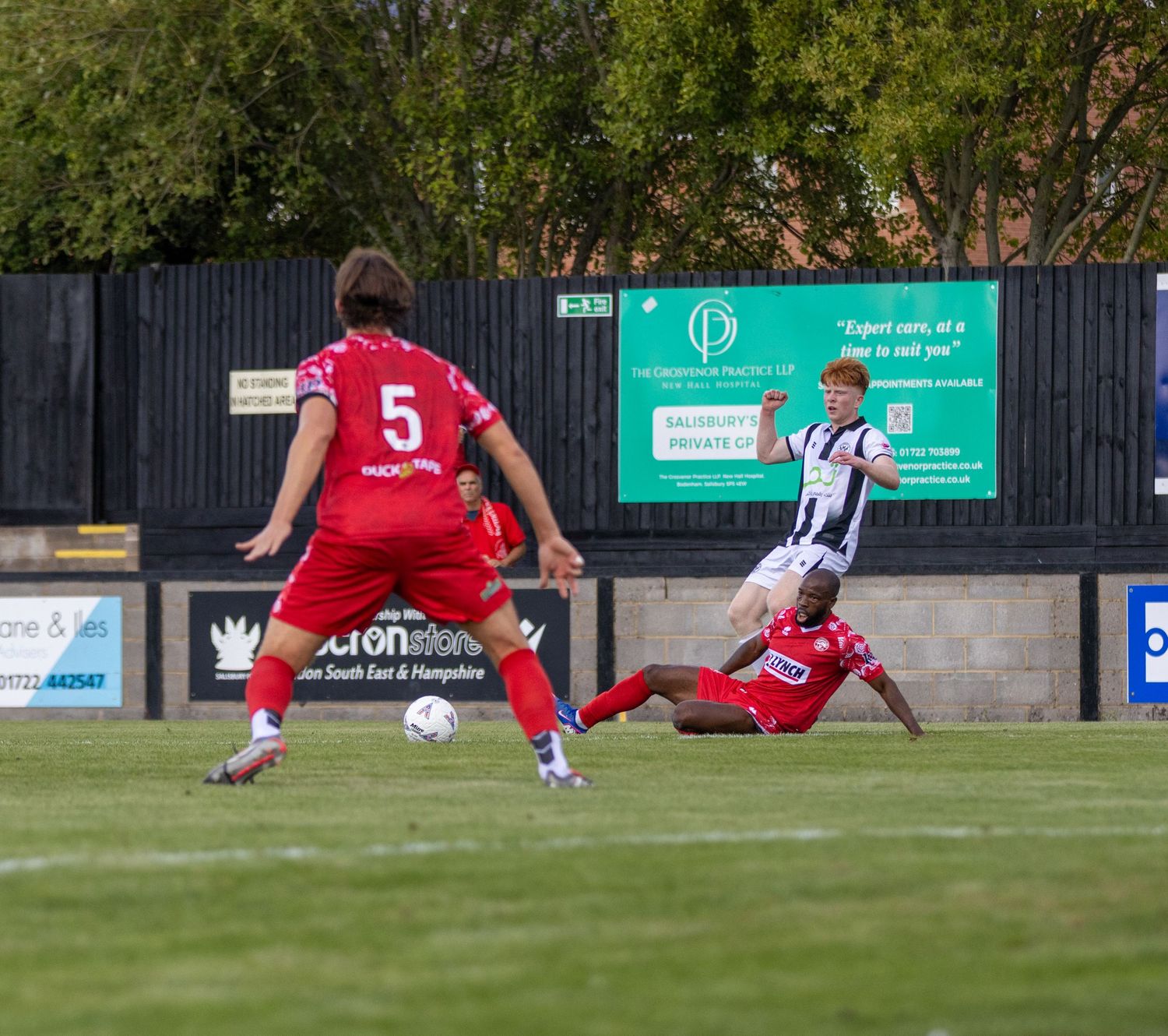 A sliding tackle challenges a black-and-white kitted player as number 5 in red looks on during a football match.
