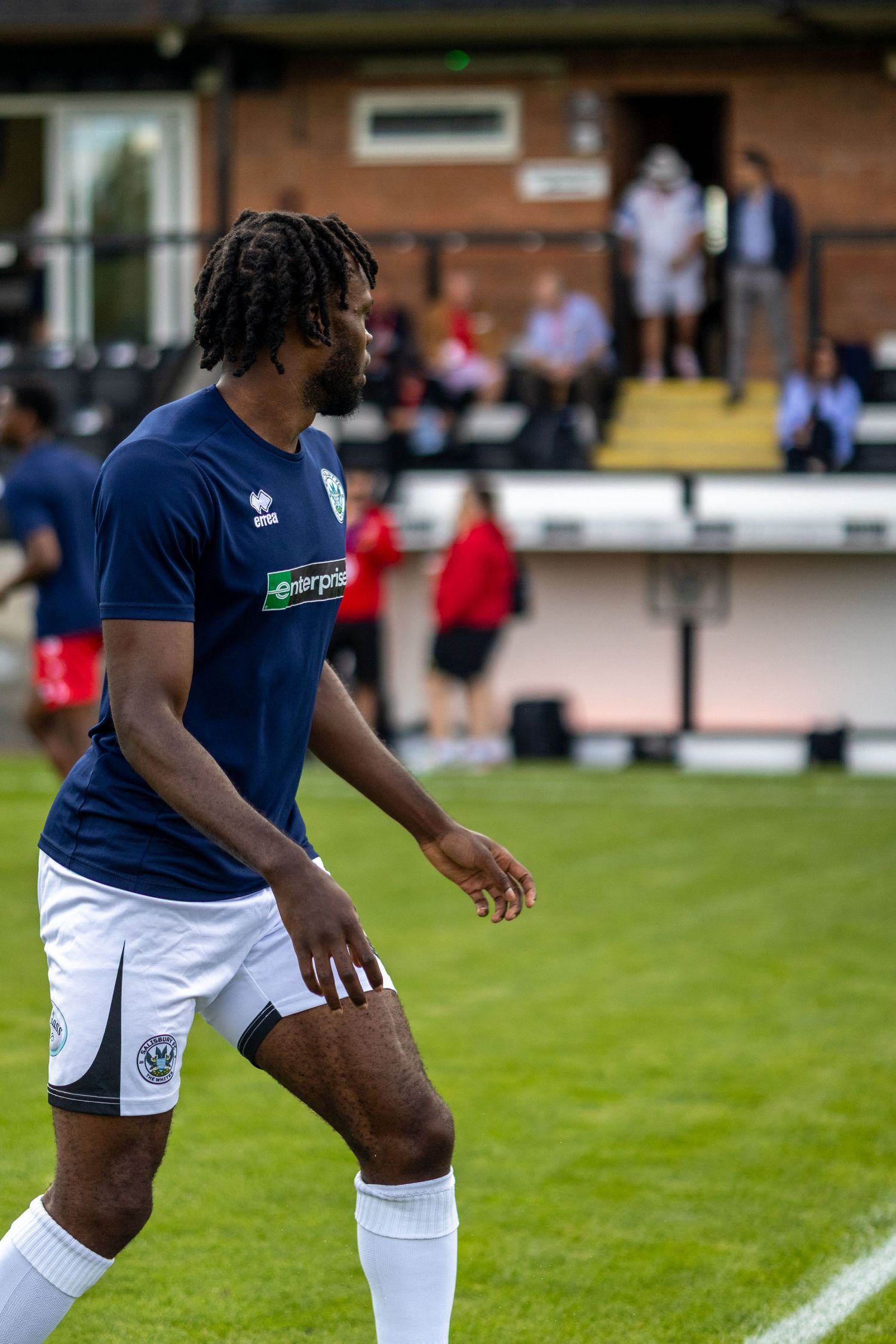 A Black male footballer in a navy Errea kit with Enterprise sponsor walks on a grass pitch during a game.