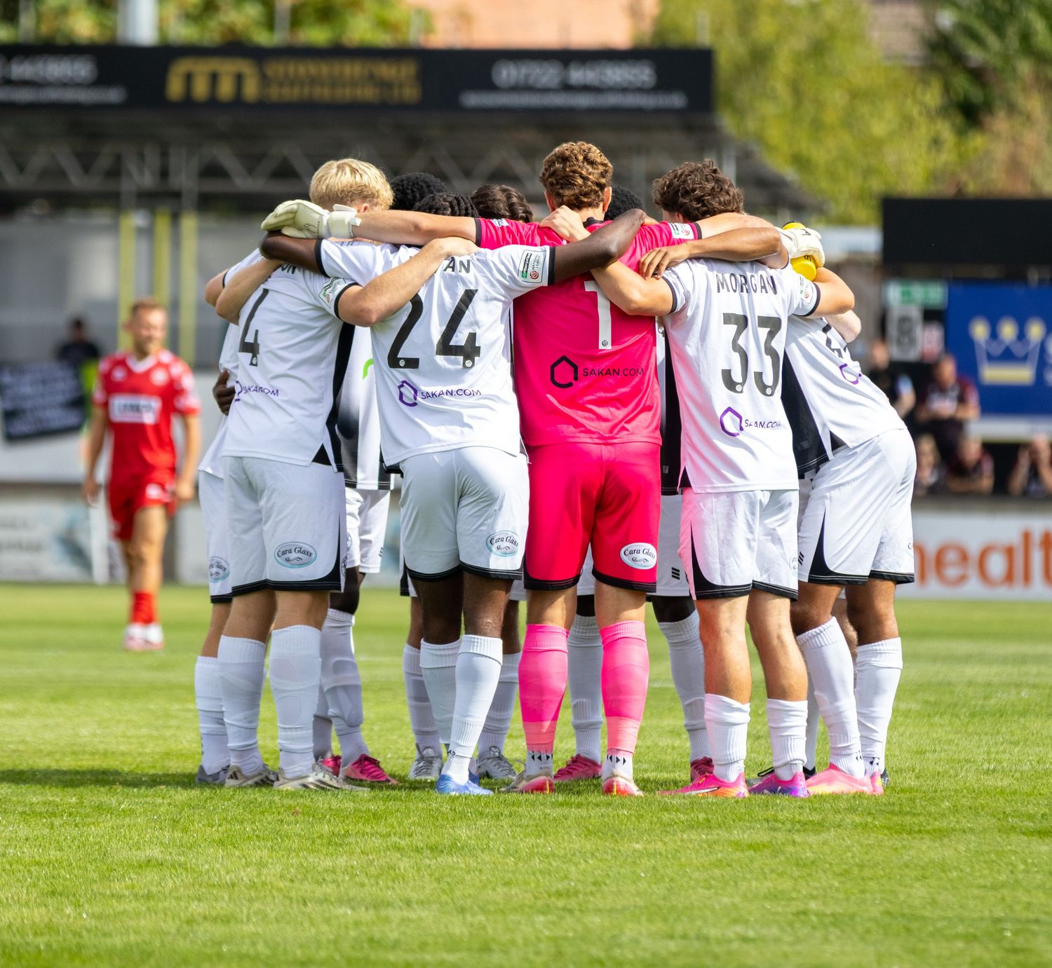 Football team in white kits and pink goalkeeper jersey huddle on sunny pitch before kickoff.