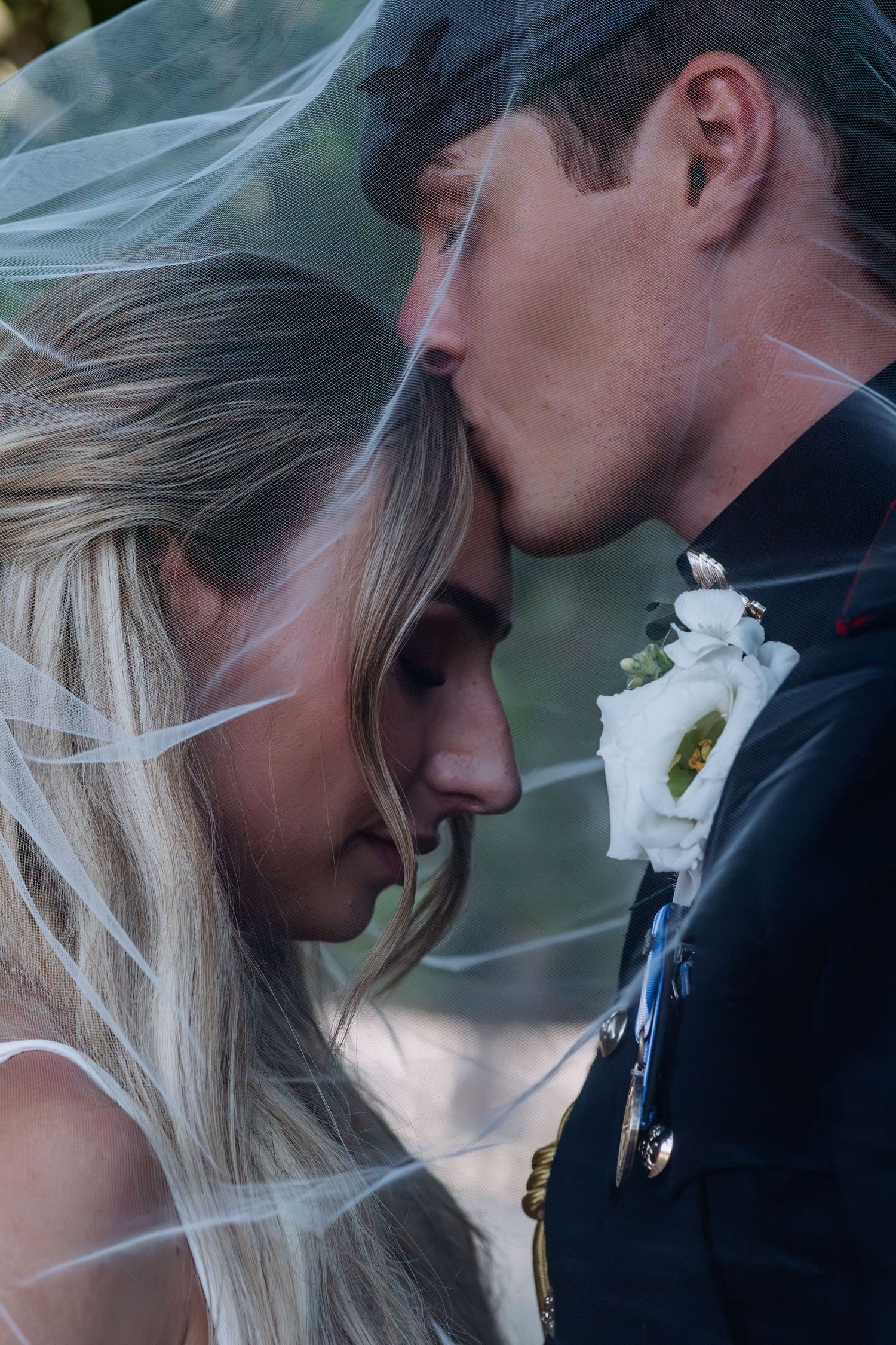 Bride and groom in military dress uniform sharing a tender forehead touch under a flowing veil.