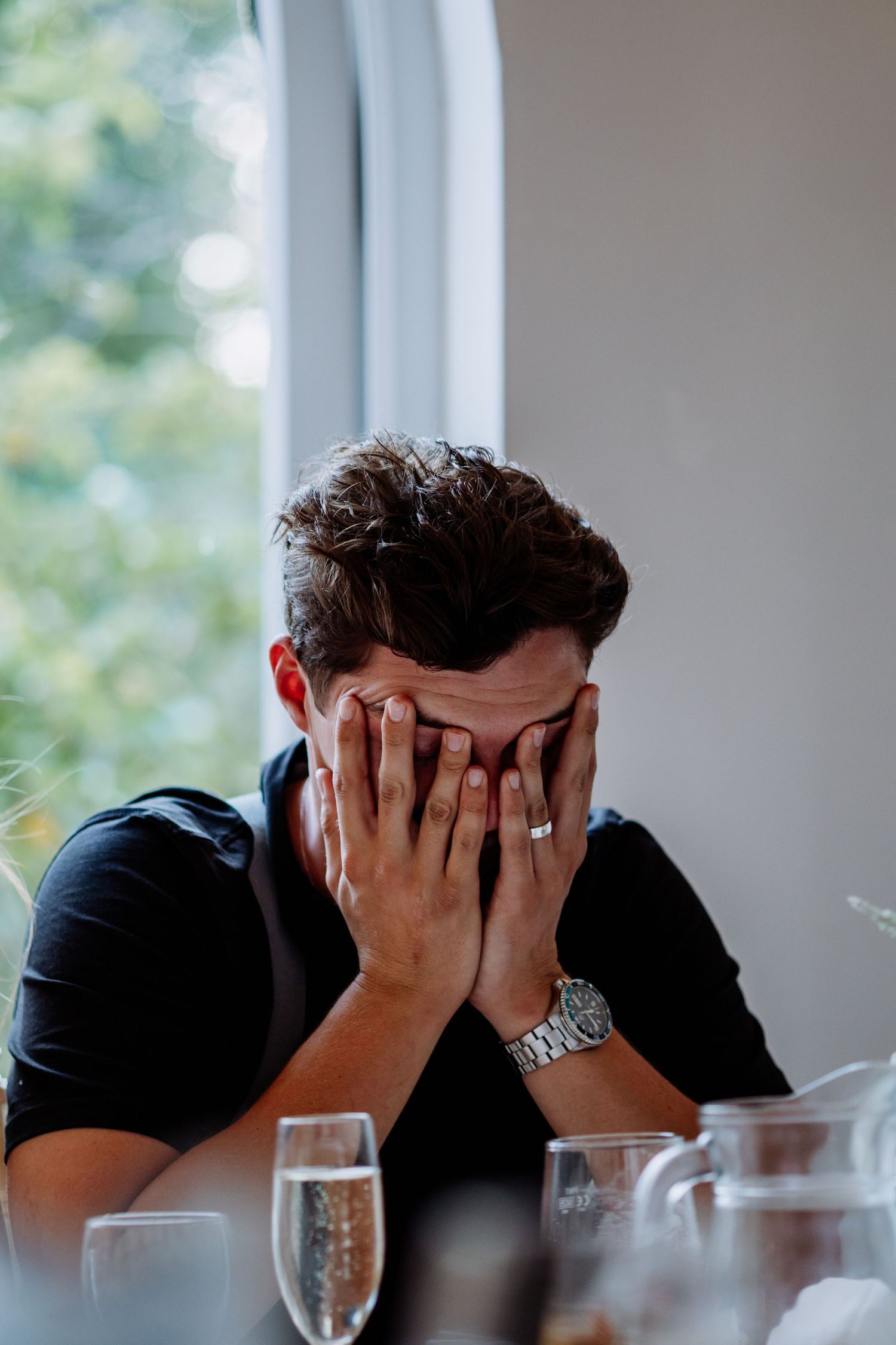 Young man in black shirt and silver watch covers face with hands emotionally at wedding reception table.