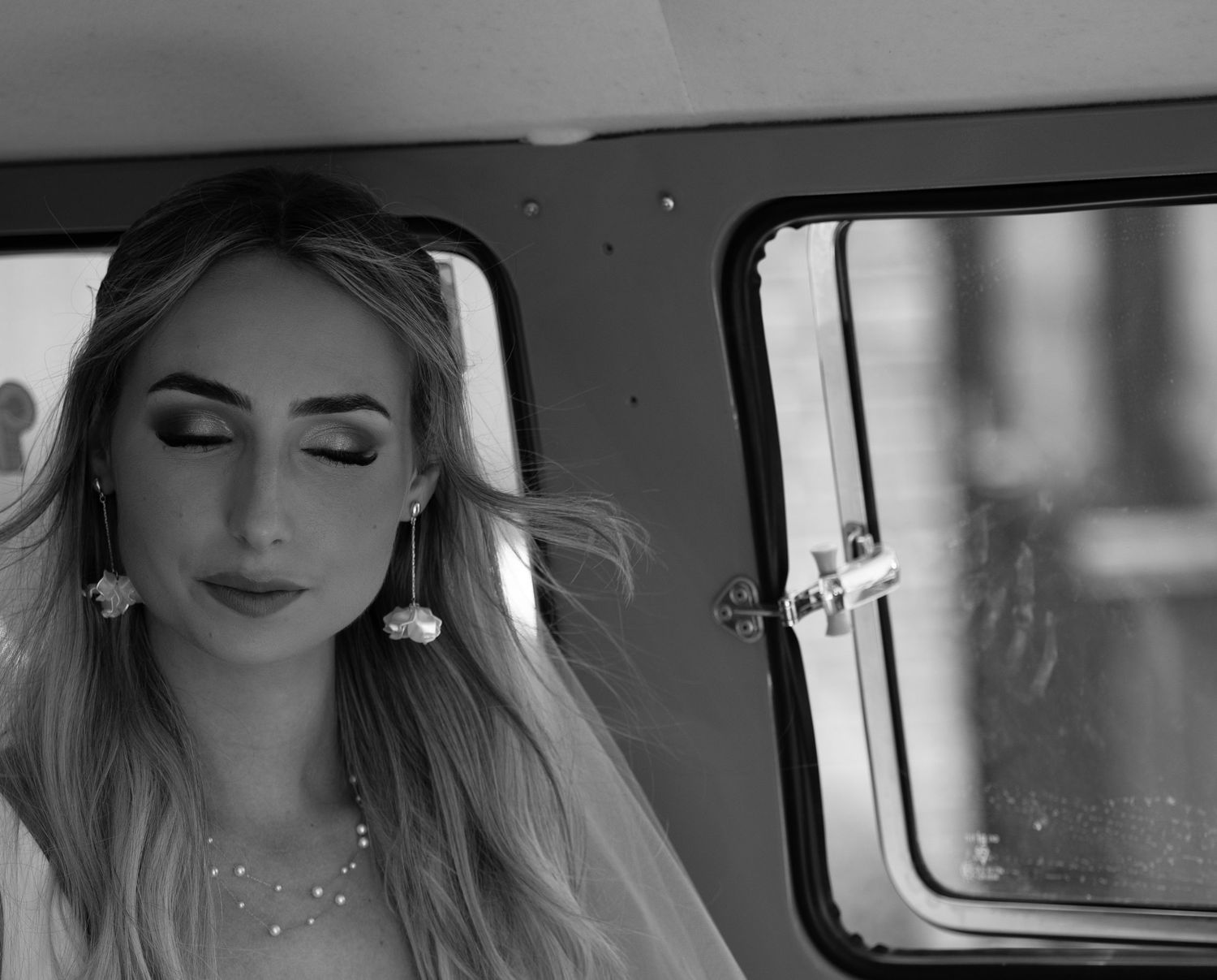 Black and white close-up of a bride wearing floral earrings and pearl necklace inside a vintage vehicle.