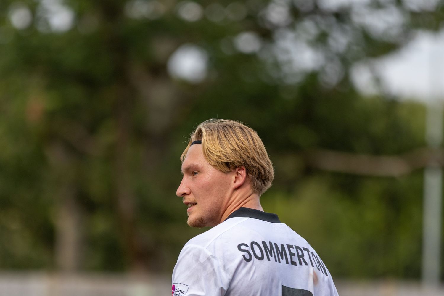 A blonde footballer in white kit with SOMMERTON on the back looks sideways on a football pitch.