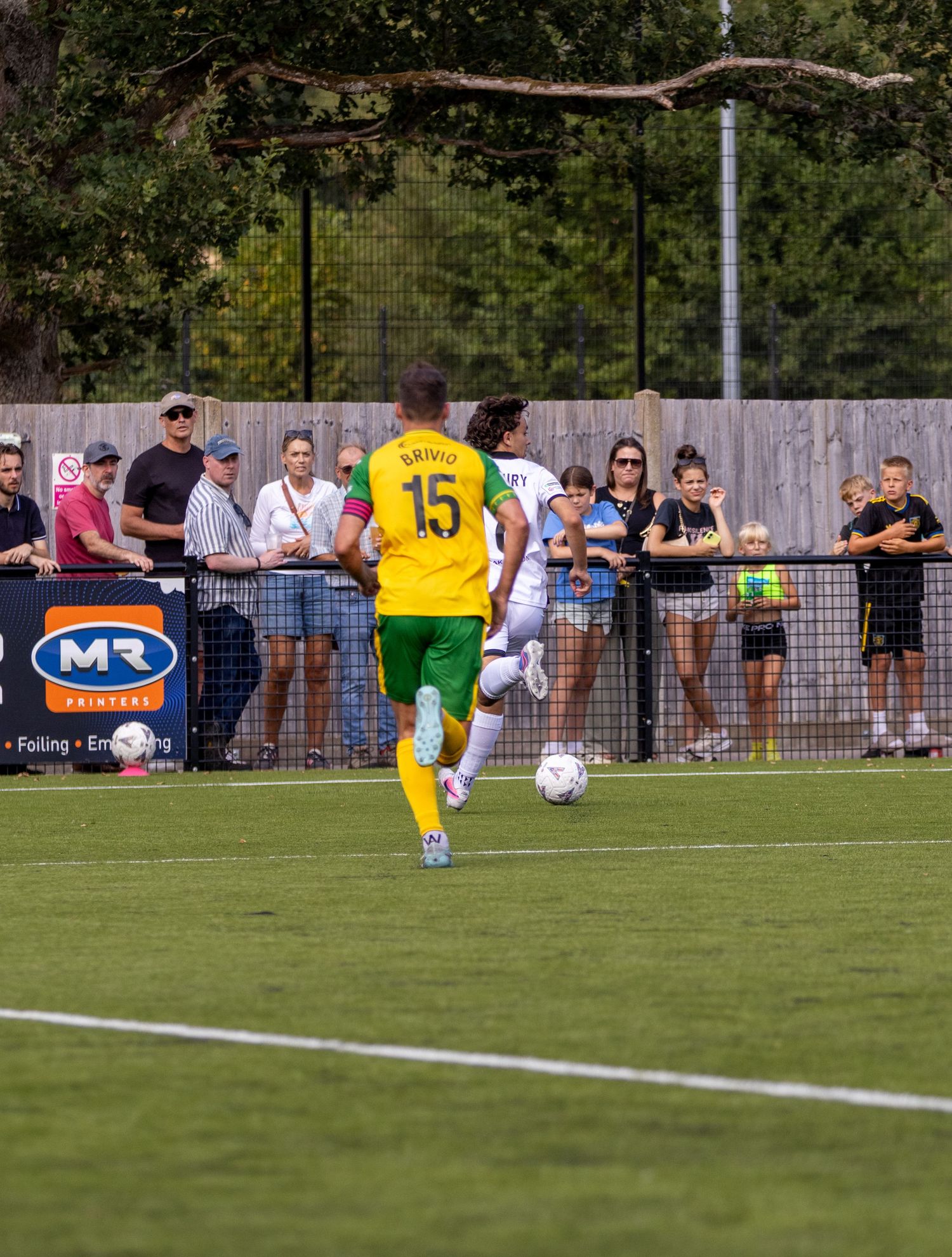 A player in yellow and green kit number 15 dribbles the ball along the sideline watched by spectators.