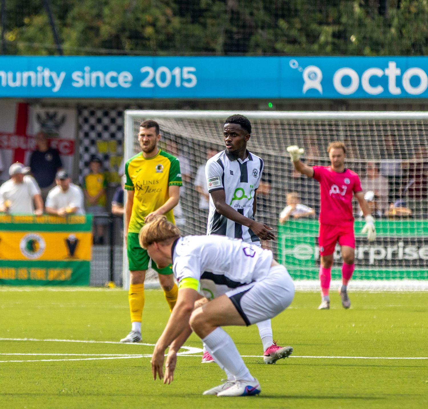 Football player in white kit kneels on pitch while teammates defend goal during match.
