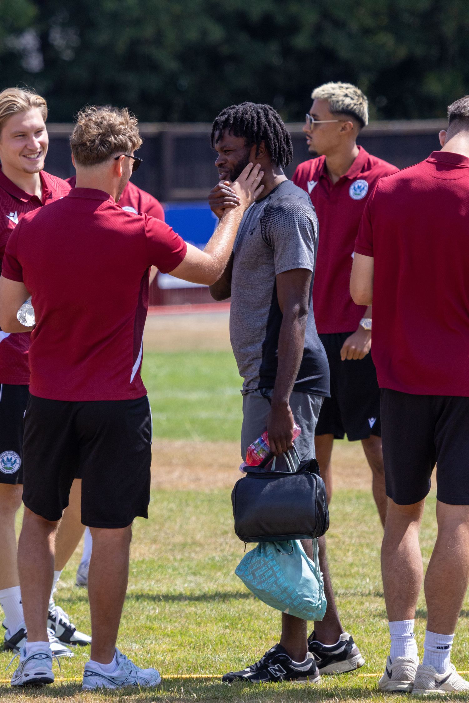 Football players in red training kit greeting dreadlocked player on sunny outdoor training ground.