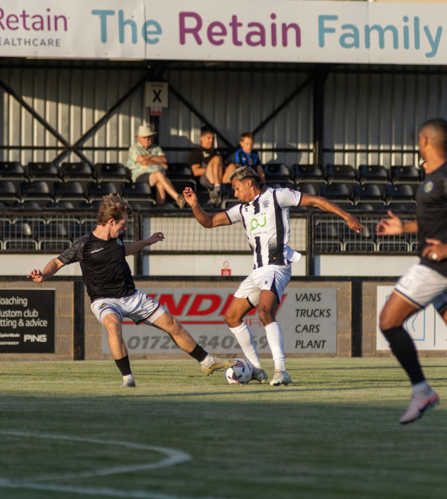 Two soccer players compete for the ball during a match at a stadium with Retain Healthcare advertising boards.