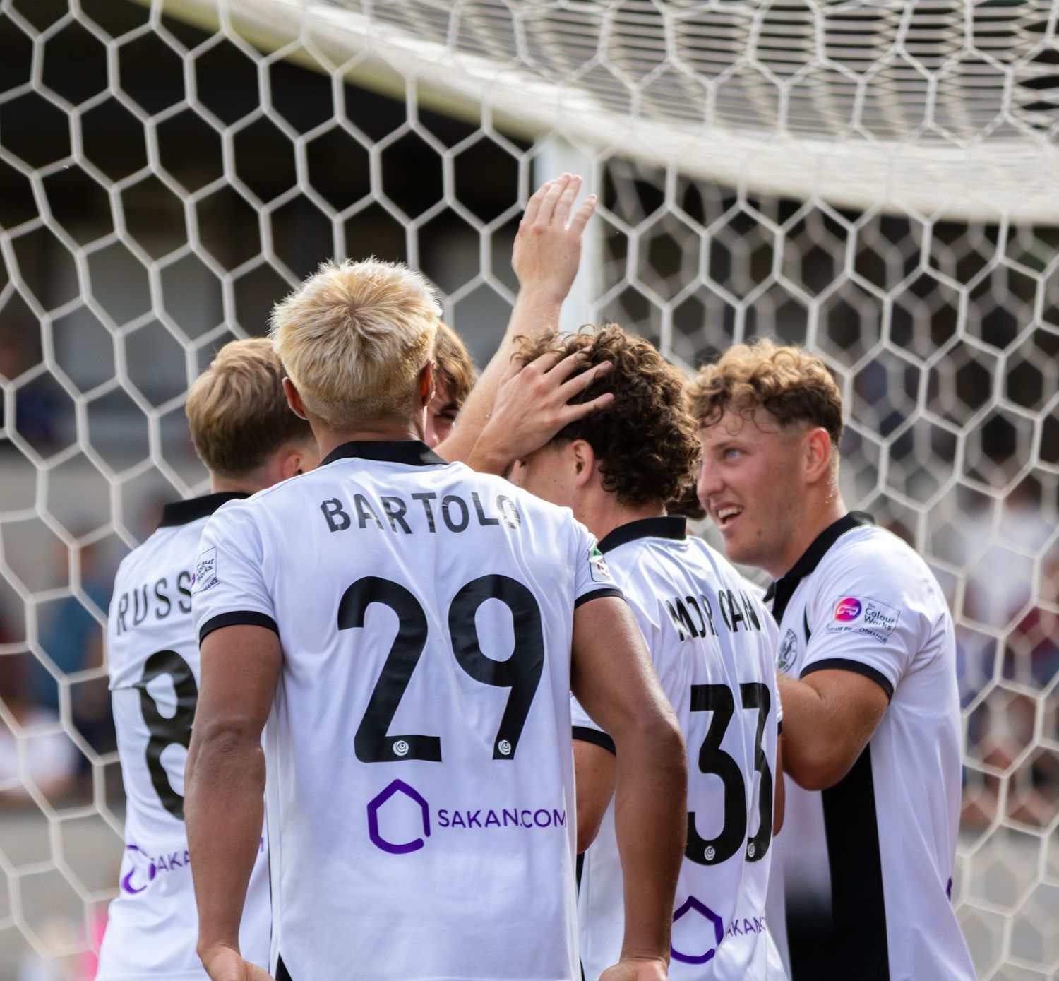 Soccer team celebrates in white uniforms, featuring players Bartoli 29 and number 33 near goal.