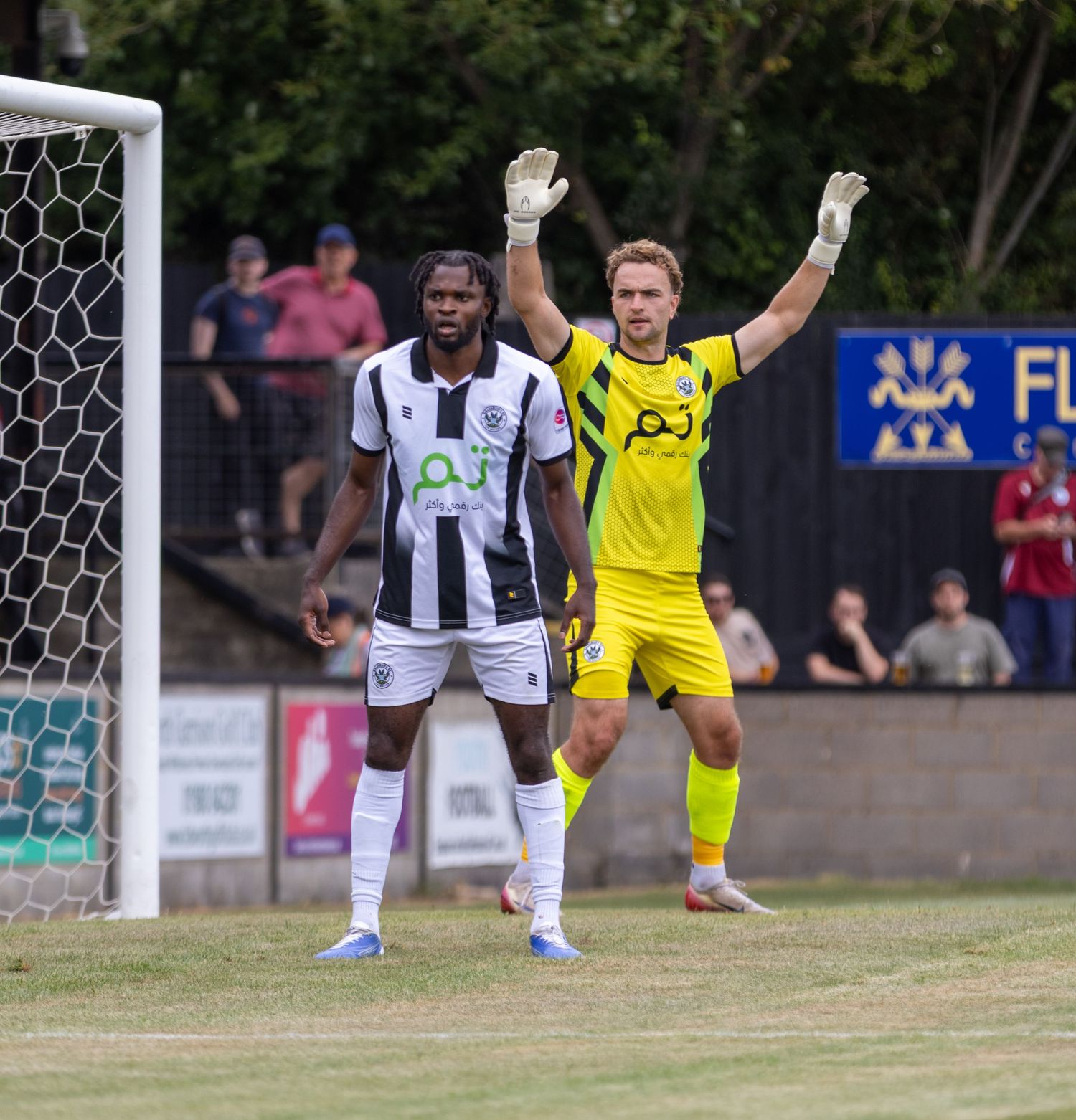A Black footballer and yellow-kitted goalkeeper stand near goal during a football match.