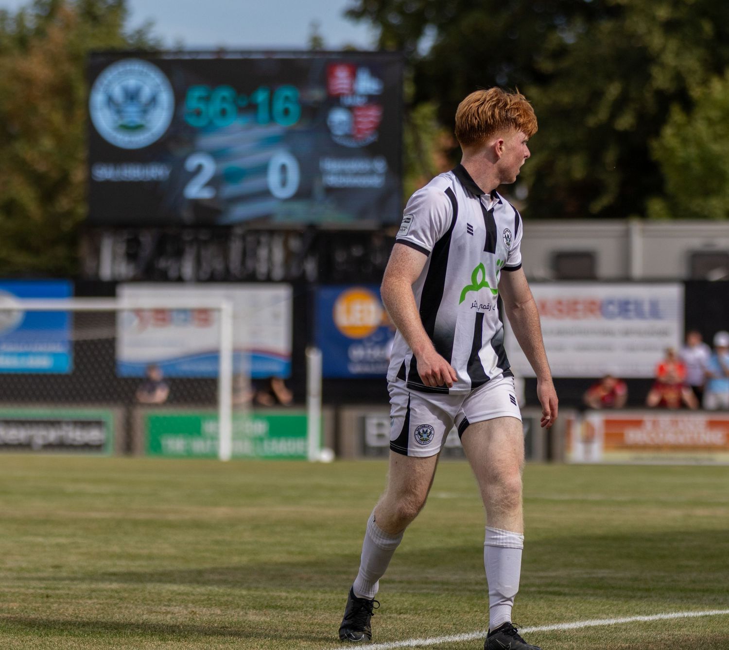 A red-haired footballer in black and white strip walks on the pitch with a 2-0 scoreboard behind.