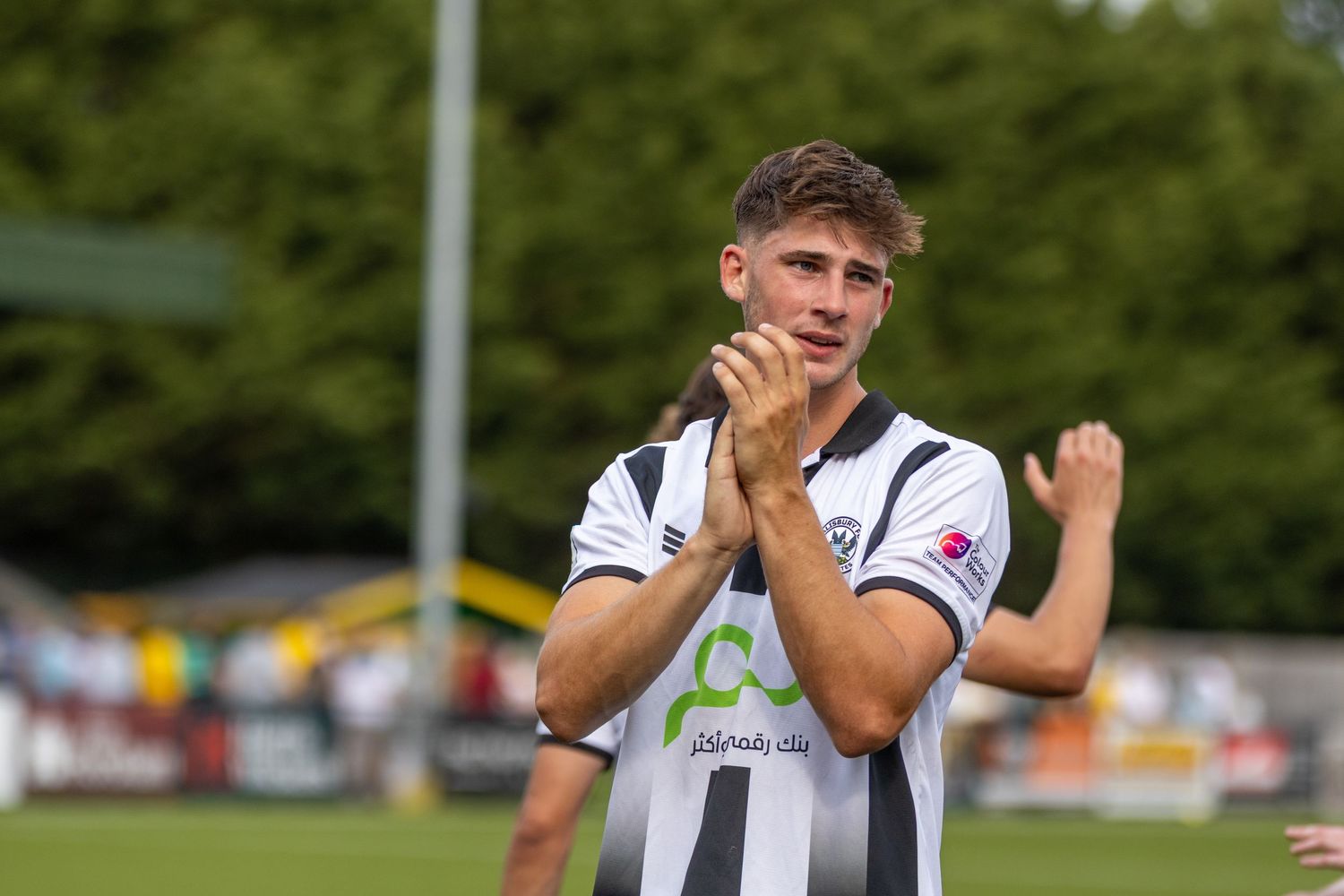 A young male footballer in black and white striped kit claps fans after a sunny football match.