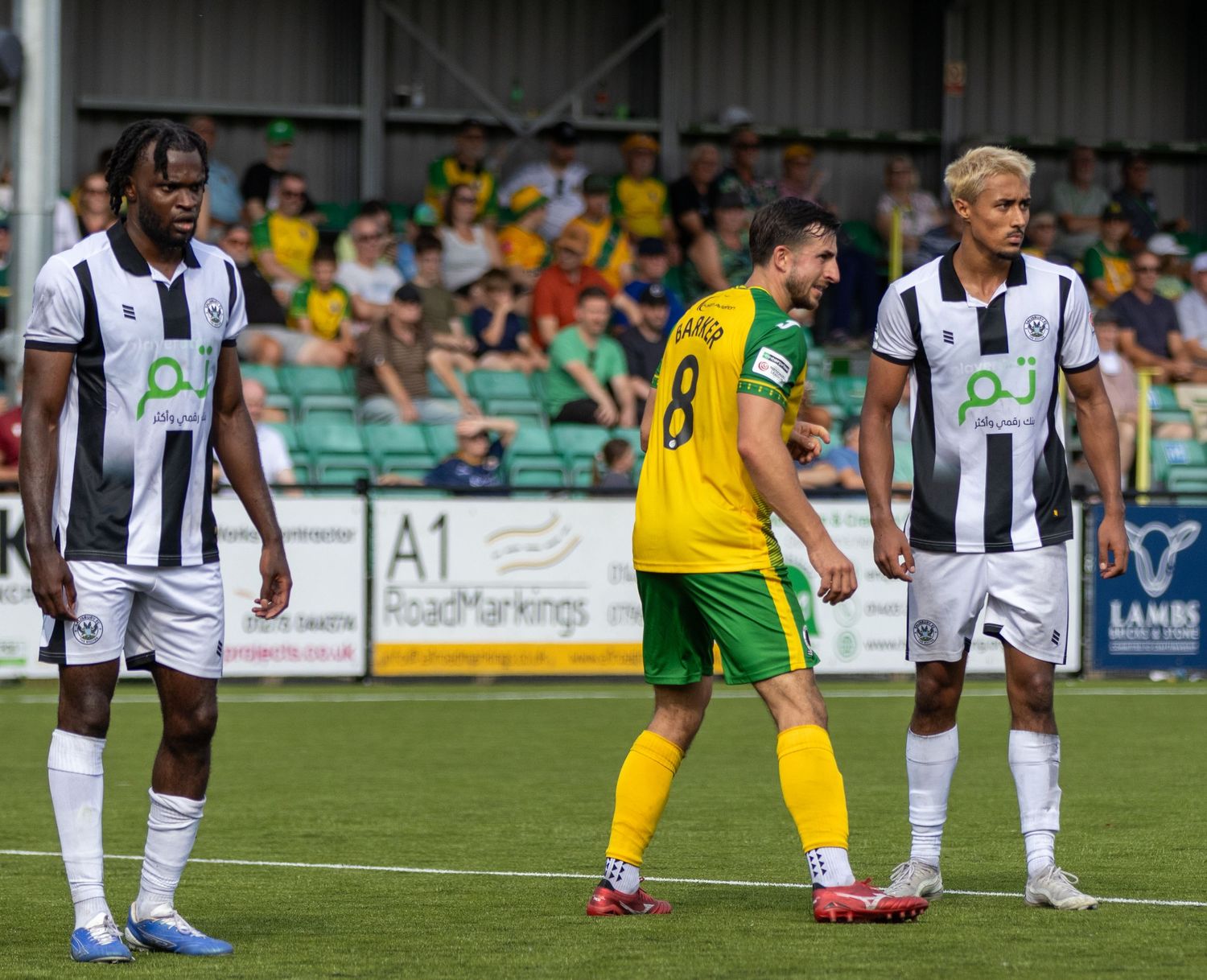 Two white kit players flank yellow kit player Barker number 8 on football pitch with fans.