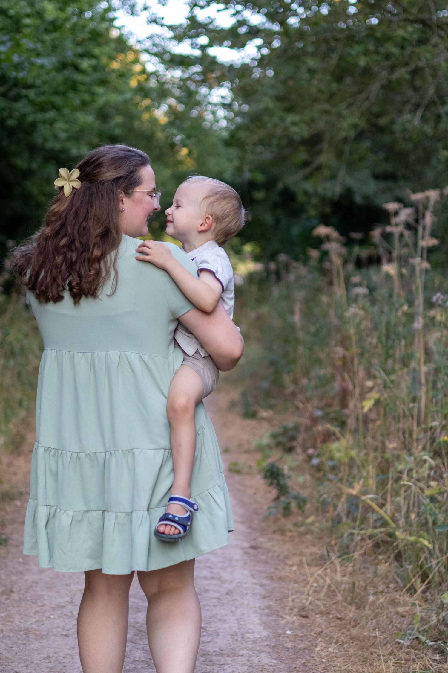 A brunette mother carrying her smiling toddler on a tree-lined dirt path in a sage green tiered dress.