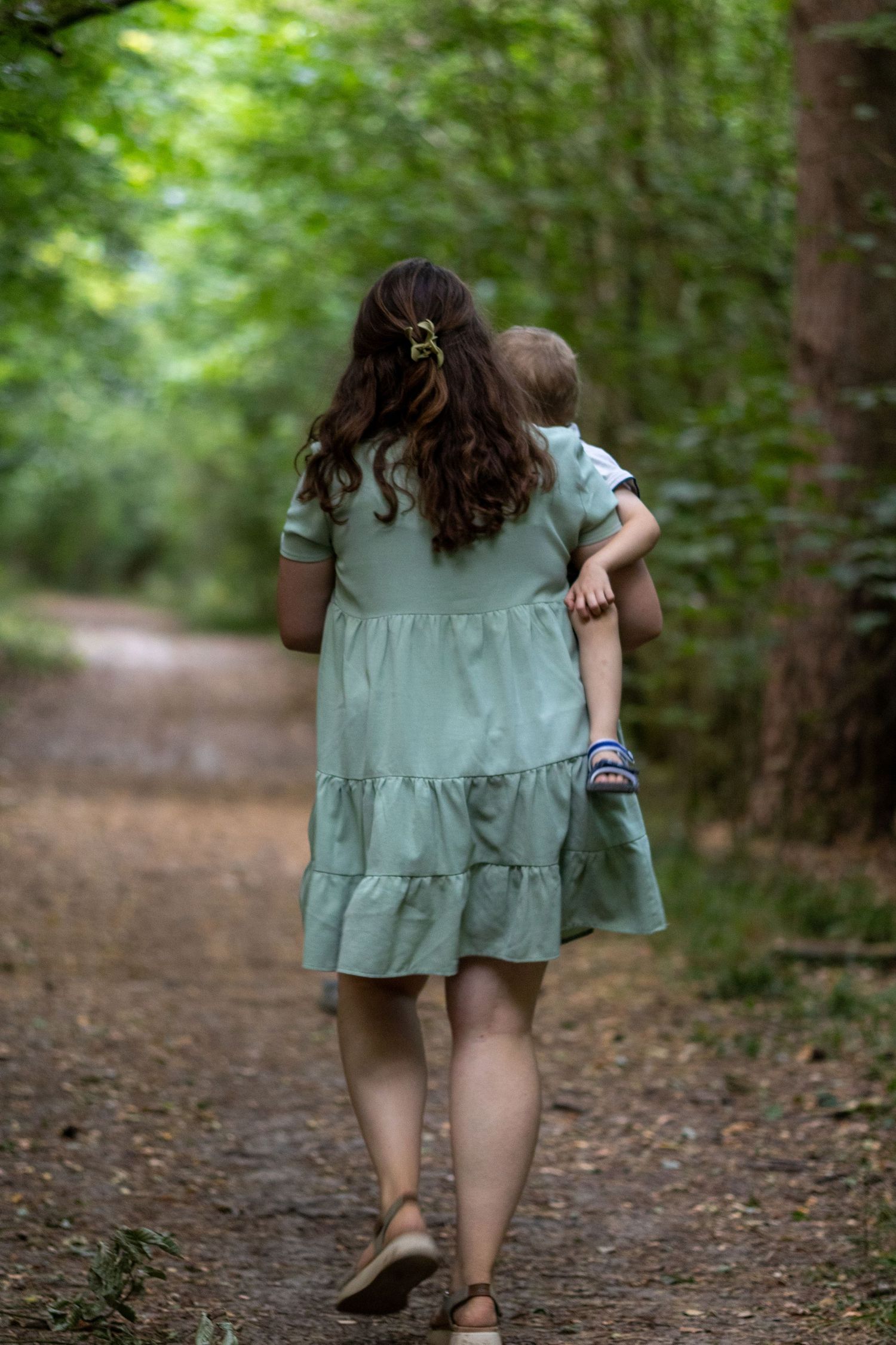 Rear view of mother in mint dress carrying toddler along a forest path.