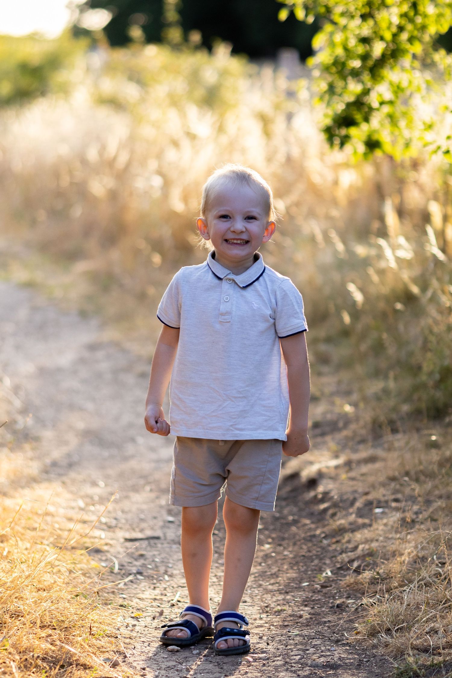 A smiling young blonde boy in a white polo and khaki shorts stands on a sunlit rural path.