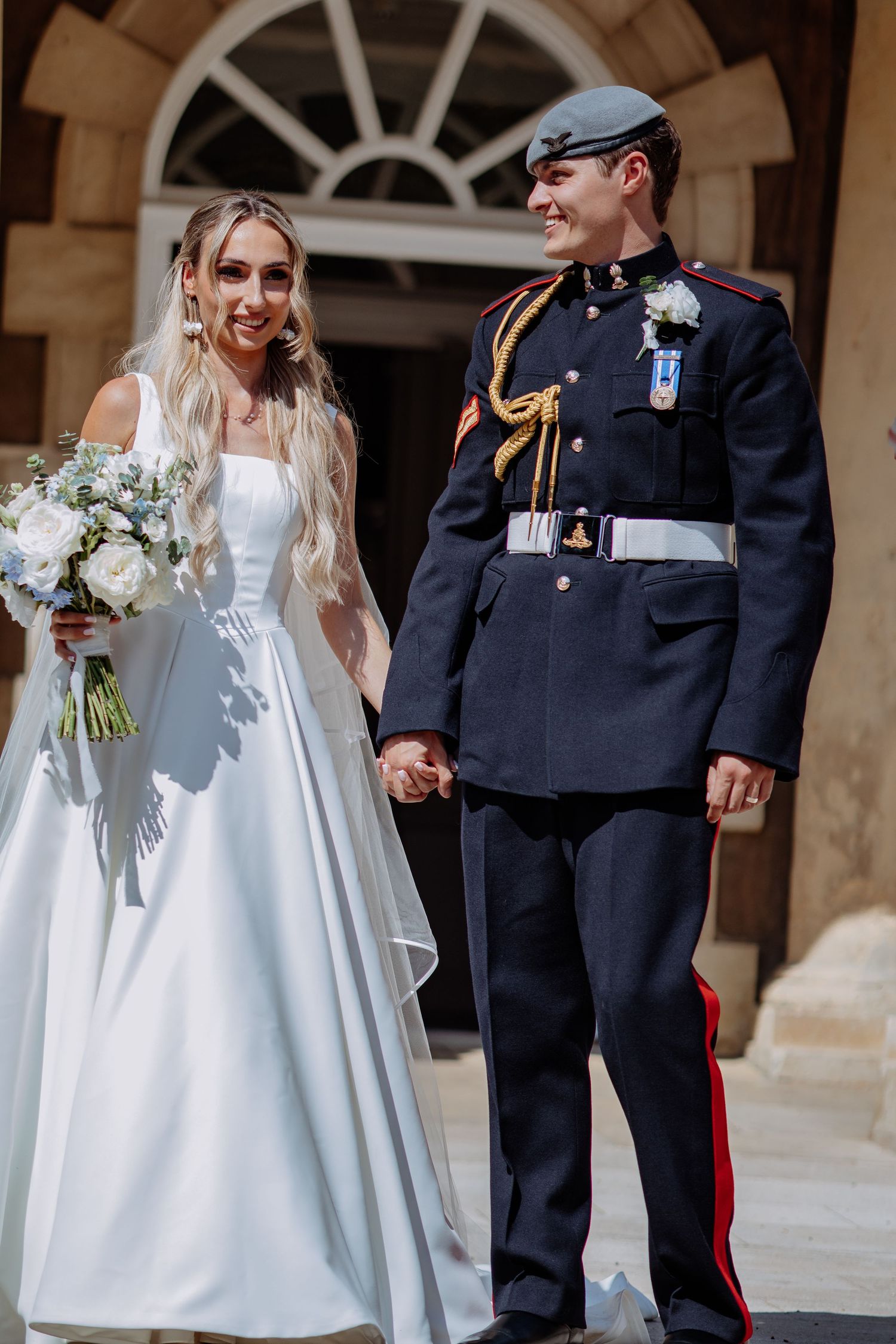 Bride and groom hold hands smiling outside stone building, groom wearing RAF ceremonial uniform with gold epaulettes.