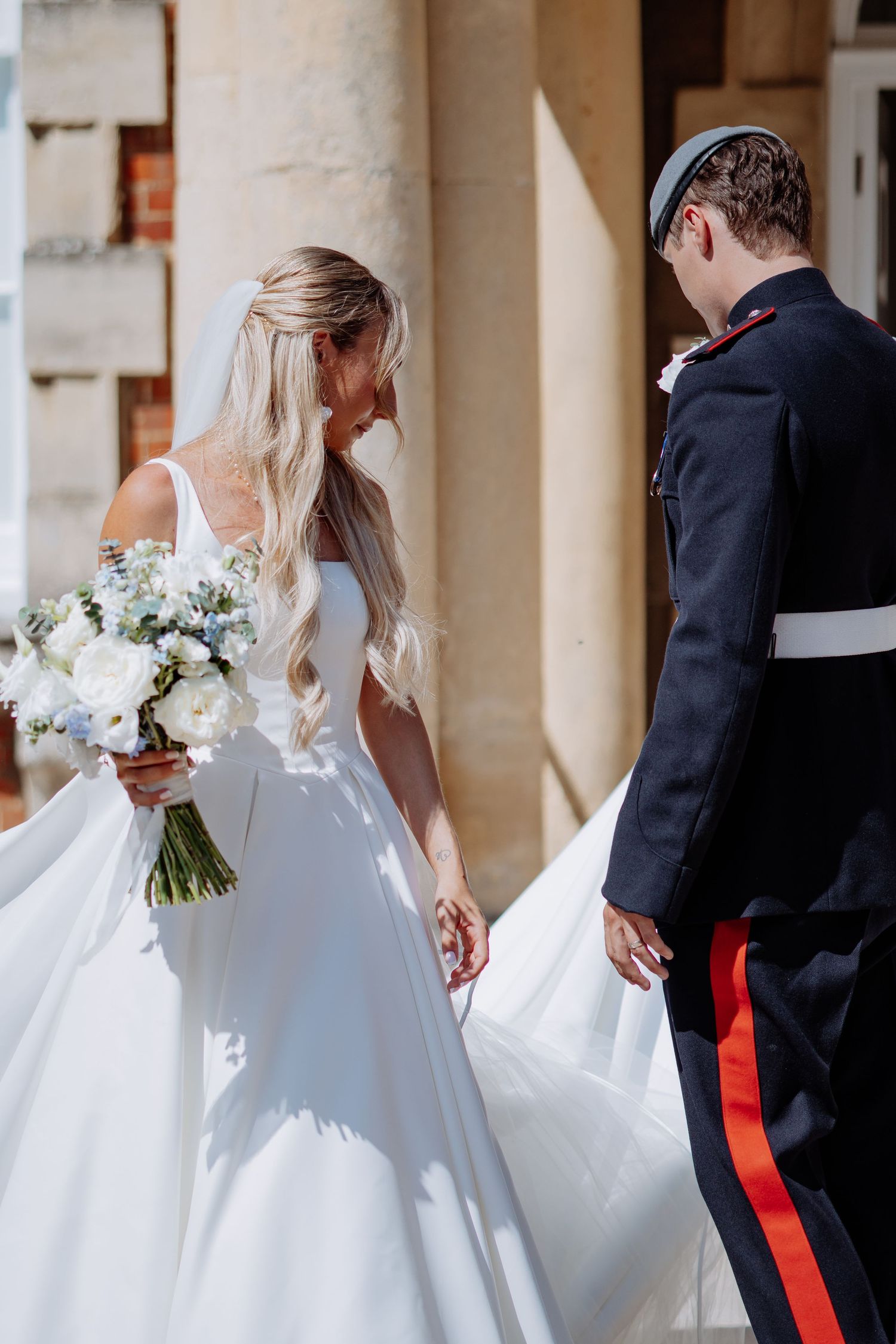 Bride in white gown holding bouquet walks with military groom in sunlight.