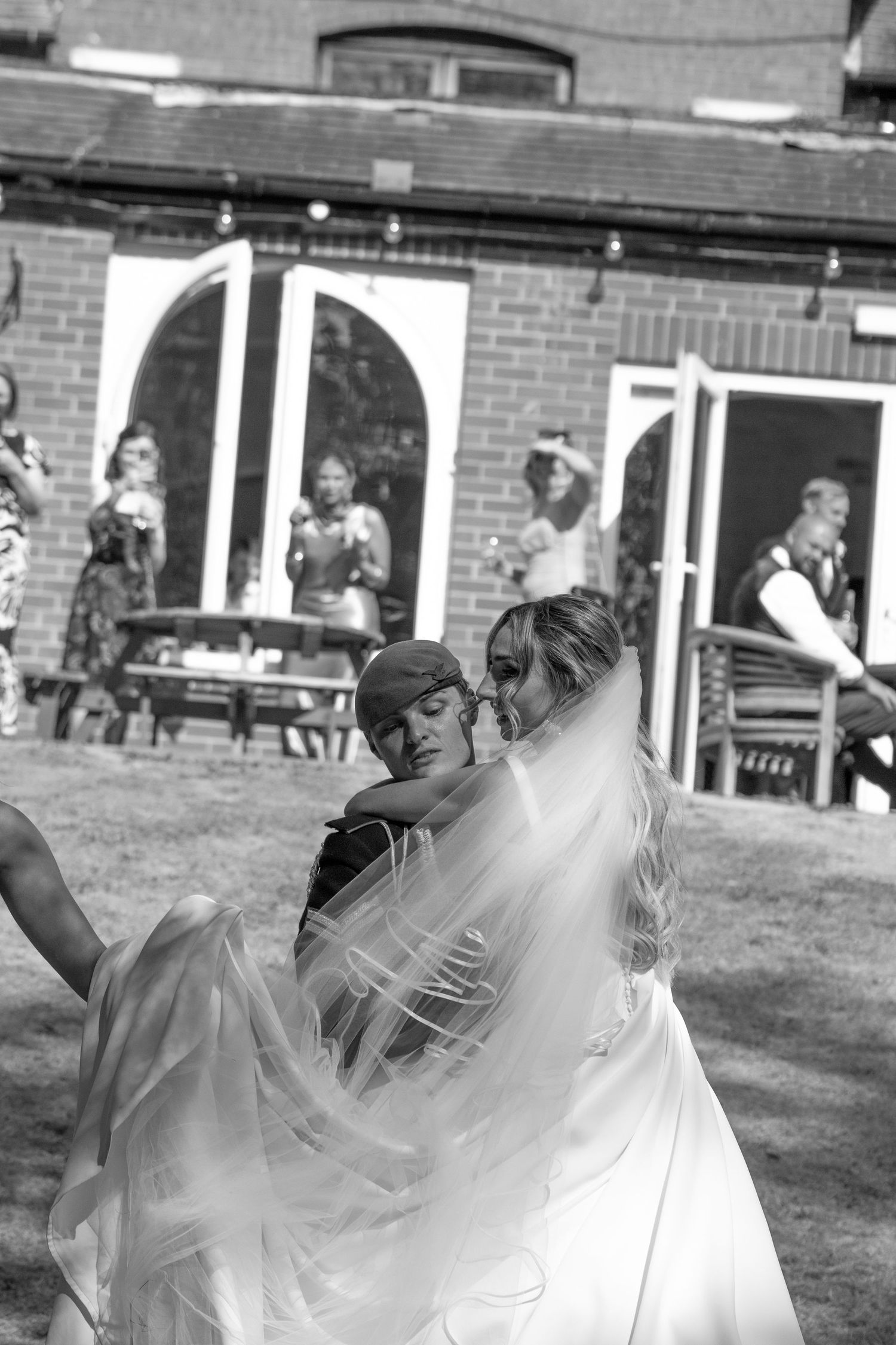 Black and white photo of bride in flowing veil embracing a guest outdoors at wedding reception.