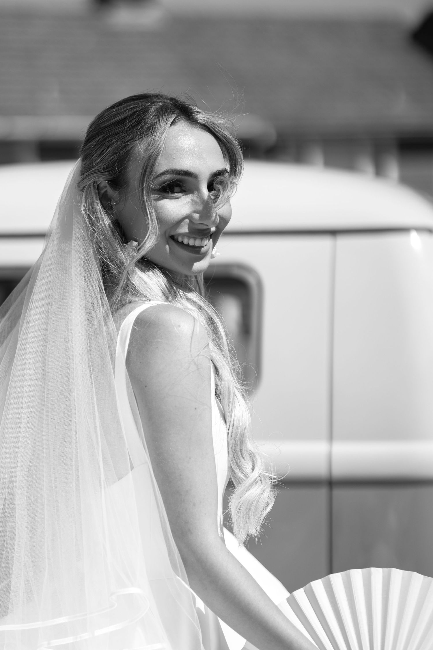 Black and white portrait of a joyful bride in veil and white dress holding a decorative fan outdoors.