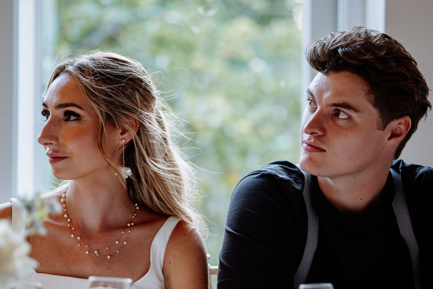 Young blonde bride in white dress and pearl necklace seated beside young man in black at wedding reception.