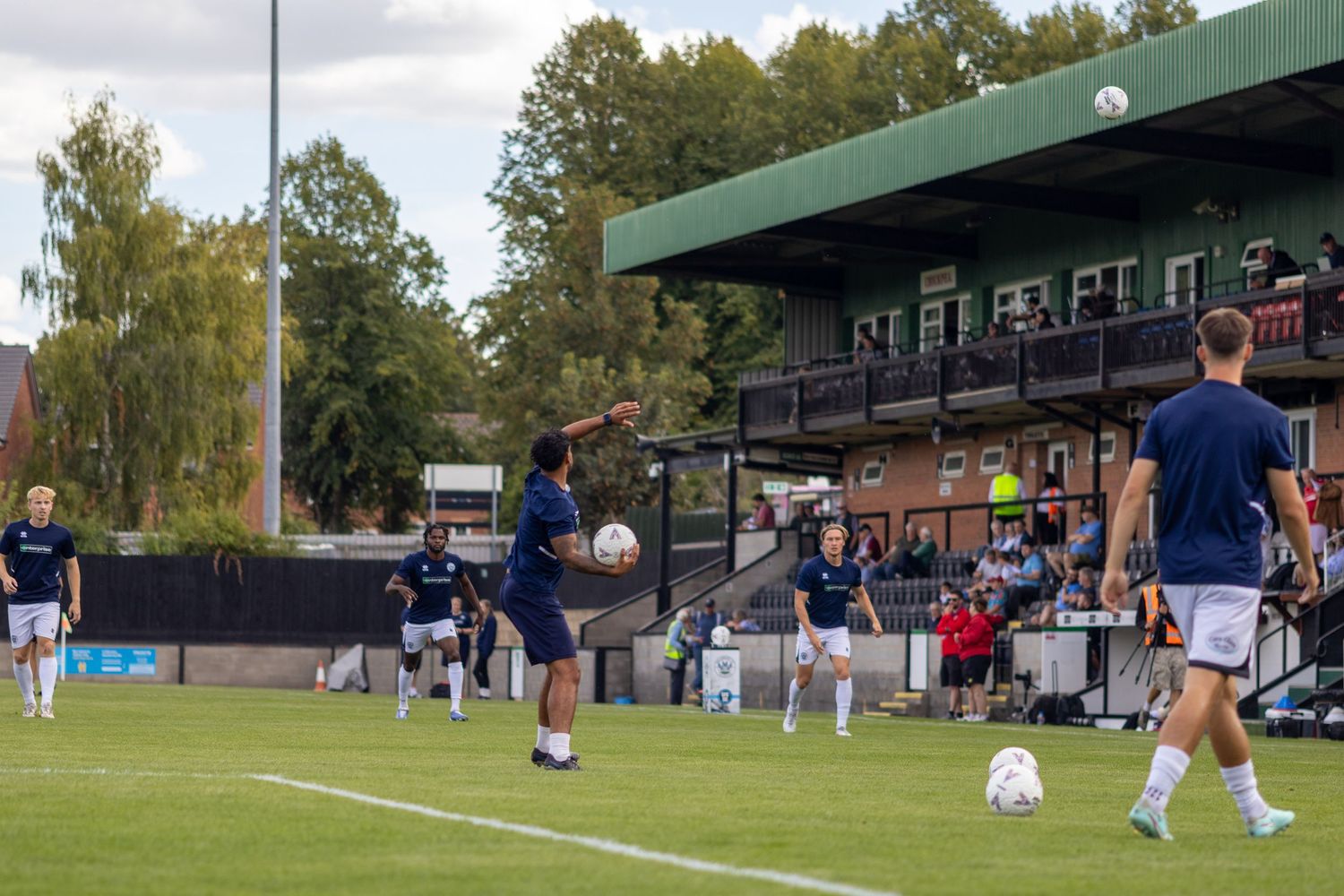 Football players in navy kits practicing passing drills on a grass pitch with spectators in the stands.