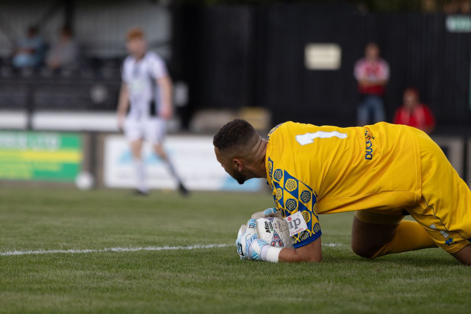 A yellow-kitted goalkeeper secures the ball on the ground during a semi-professional football match.