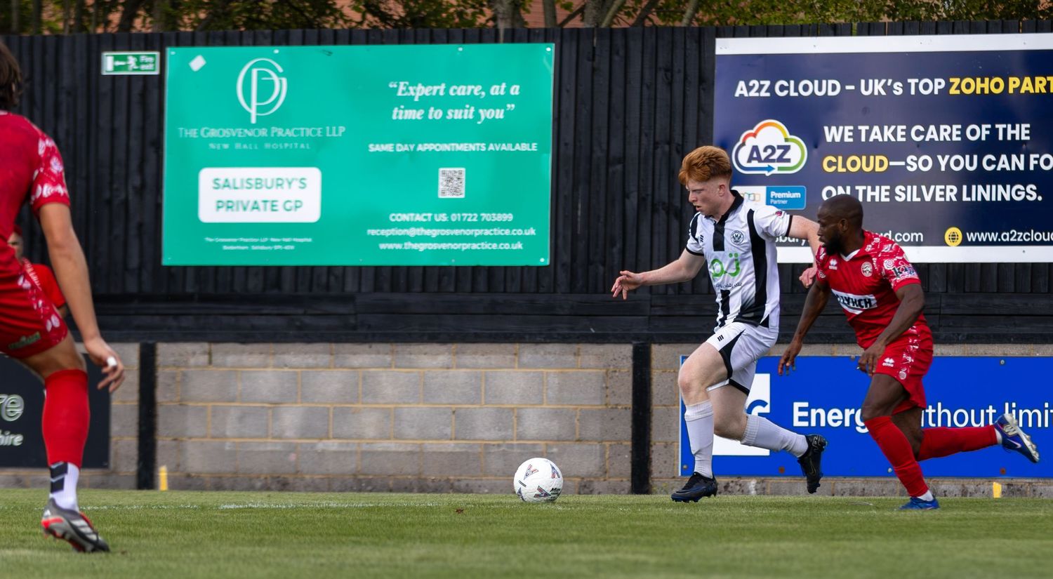 A black-and-white kitted player drives forward with the ball past a red-shirted opponent near advertising boards.