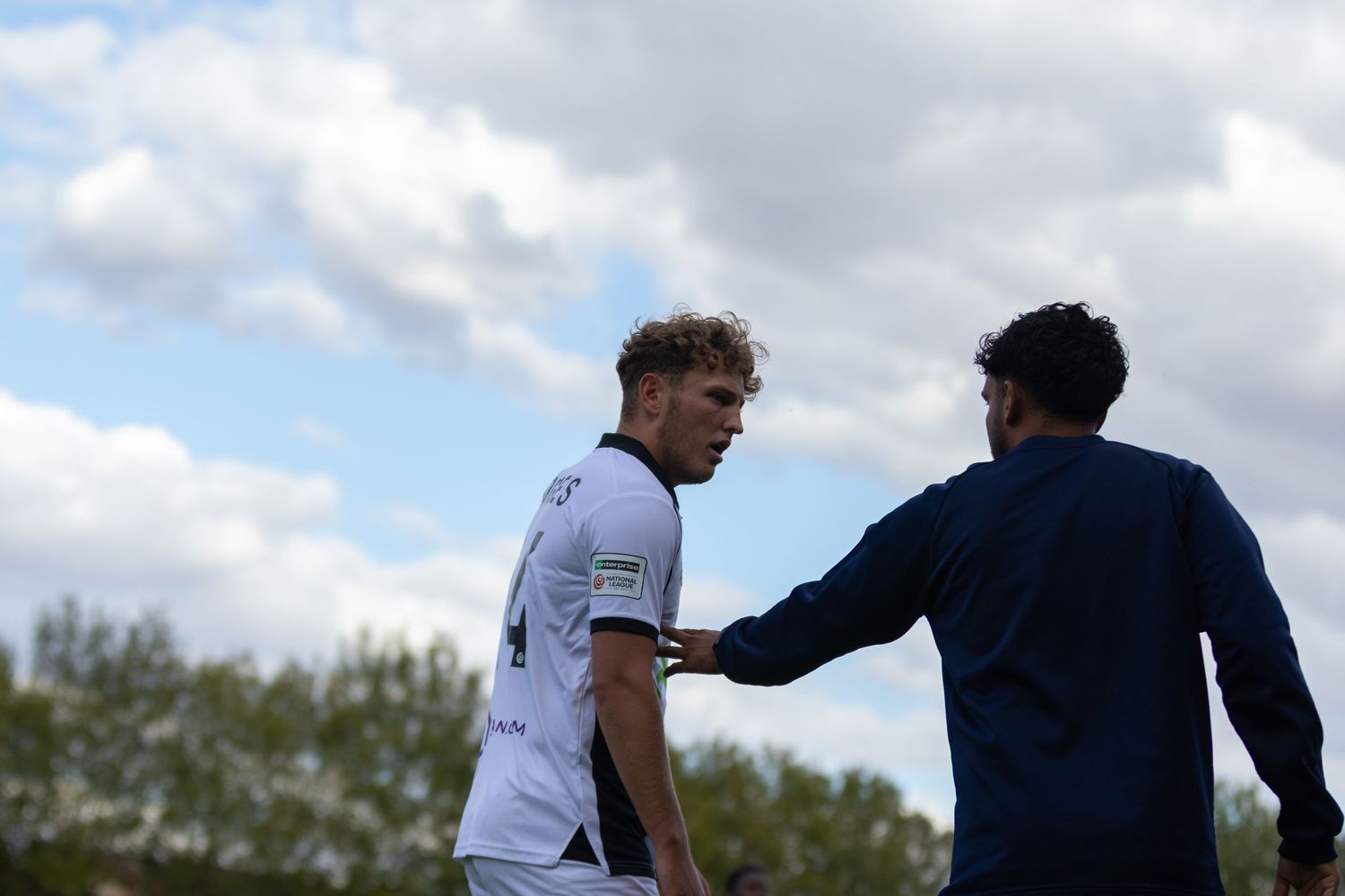 Two soccer players in white and blue uniforms discussing strategy on a green field under a cloudy sky.