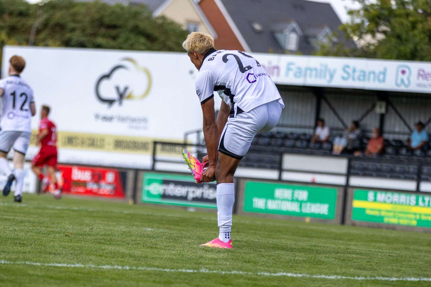 A footballer in white kit stretches on the pitch near the Retain Family Stand during a football match.