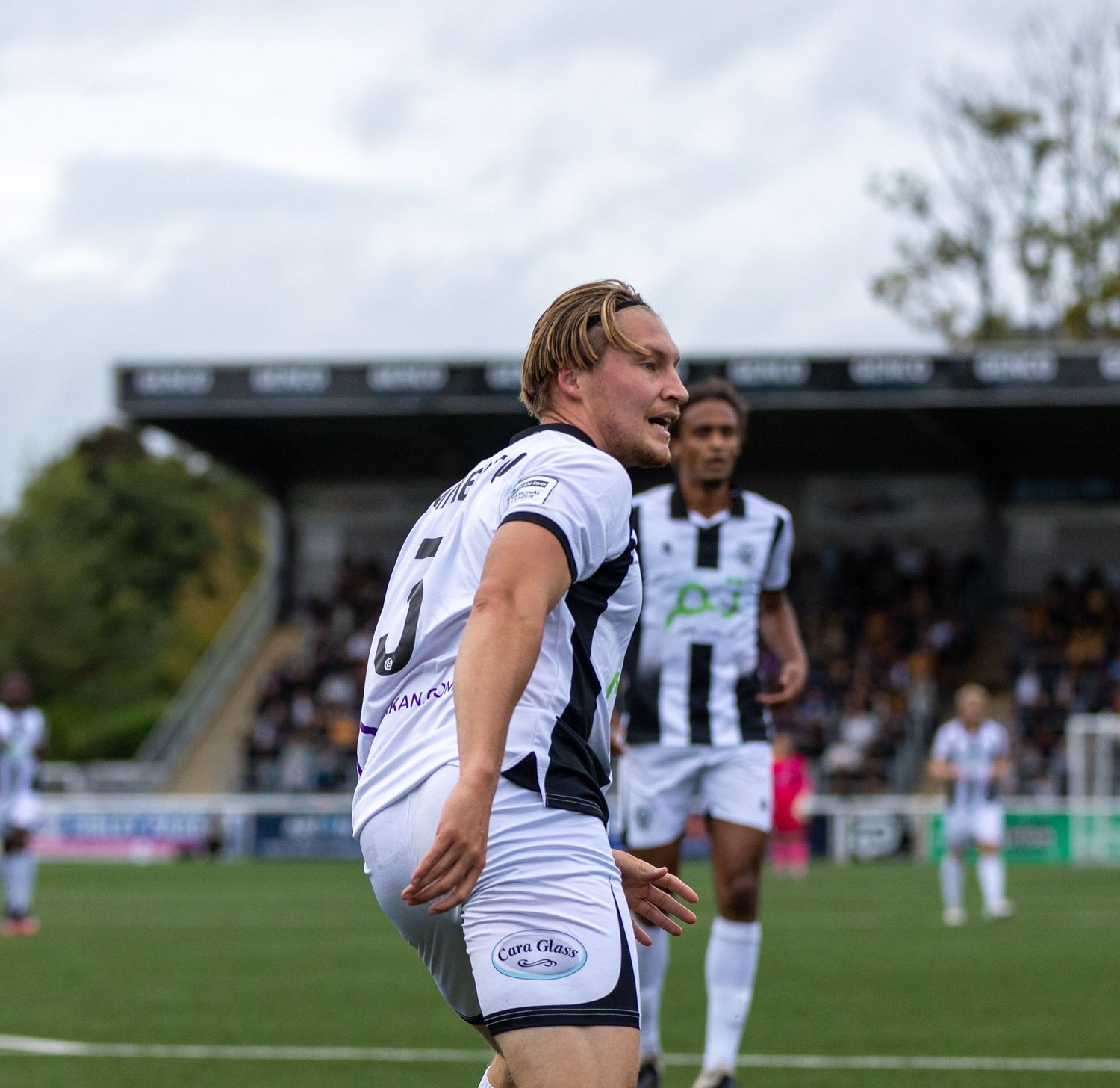 Football player in white and black kit jogging on pitch during a match, with stadium stand and teammates visible behind.
