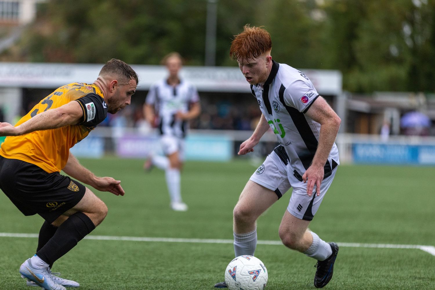 Two soccer players compete for the ball during a match, one in black and white kit, the other in yellow and black.