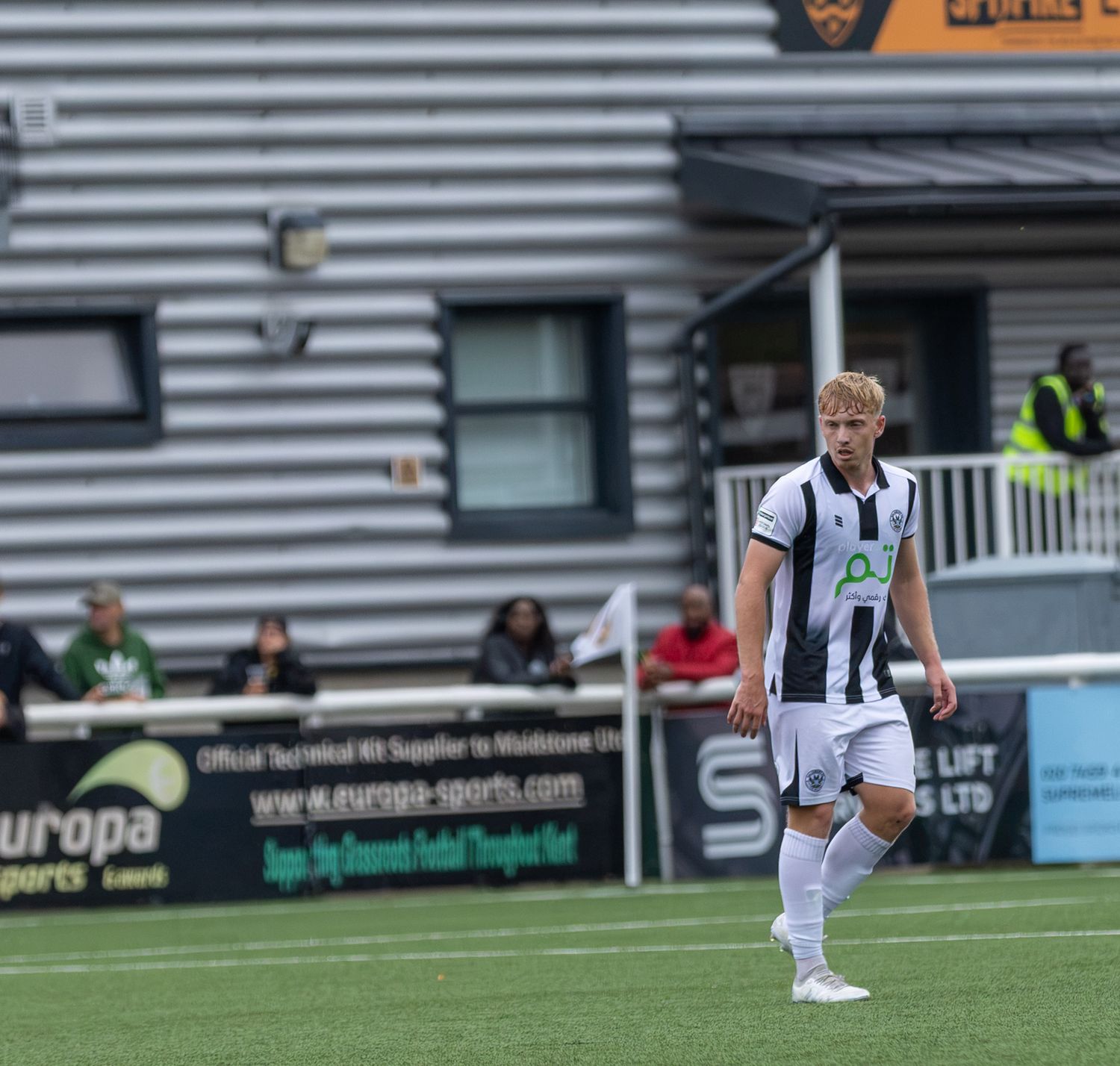 A blonde footballer in black and white kit walks on the pitch during a match at a non-league ground.