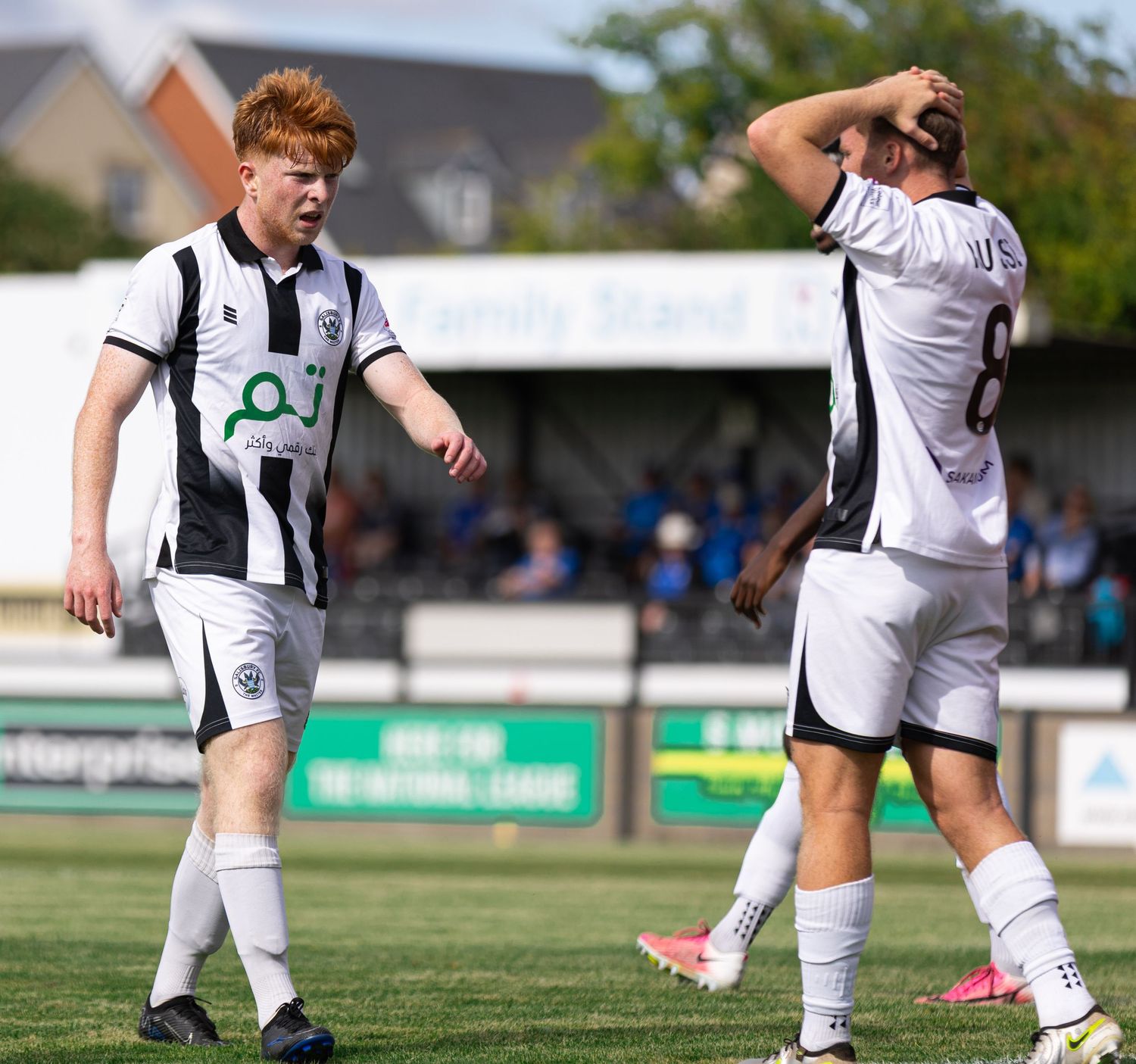 Two soccer players in black and white uniforms on a sunny match day, one with red hair walking on the pitch.