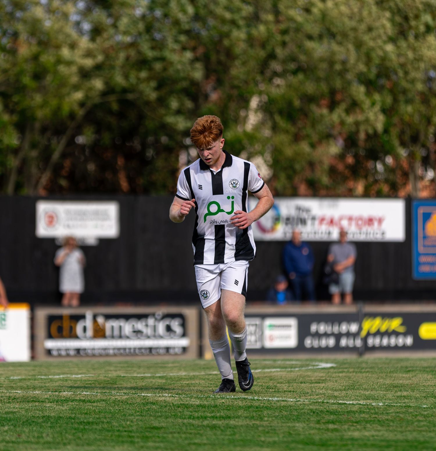 A young redhead soccer player in black and white uniform jogging on a green field during a sunny outdoor match.