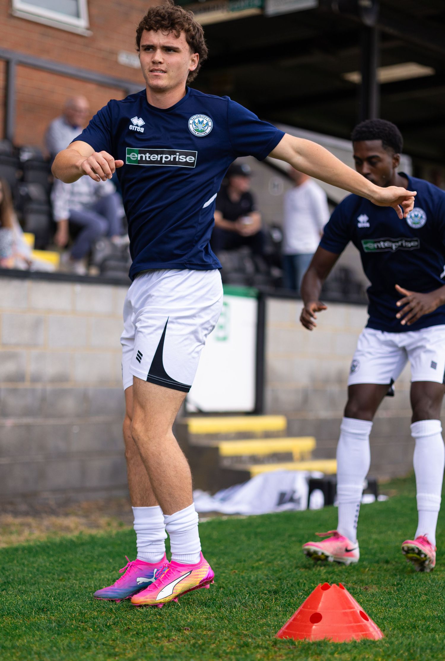 A soccer player in a navy Enterprise-sponsored jersey and white shorts warms up with stretching drills on a grass field.