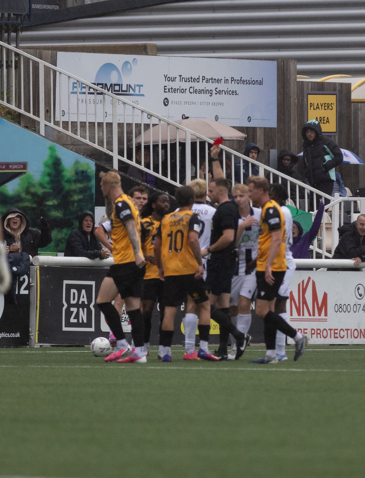 Football players in yellow and black jerseys clash on the pitch as a referee shows a red card during a match.