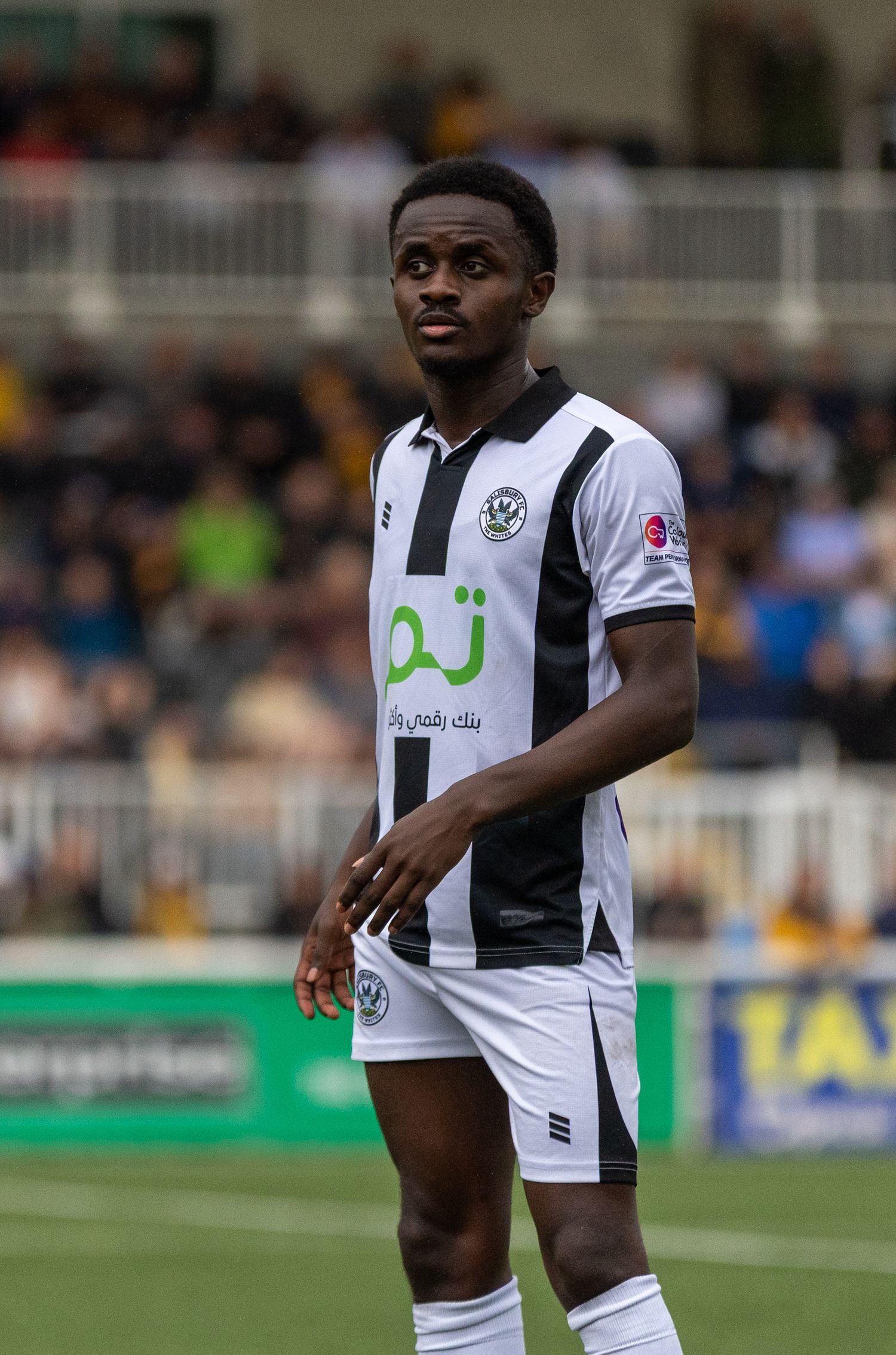 Young male soccer player in black and white striped kit with Arabic sponsor logo standing on football pitch.